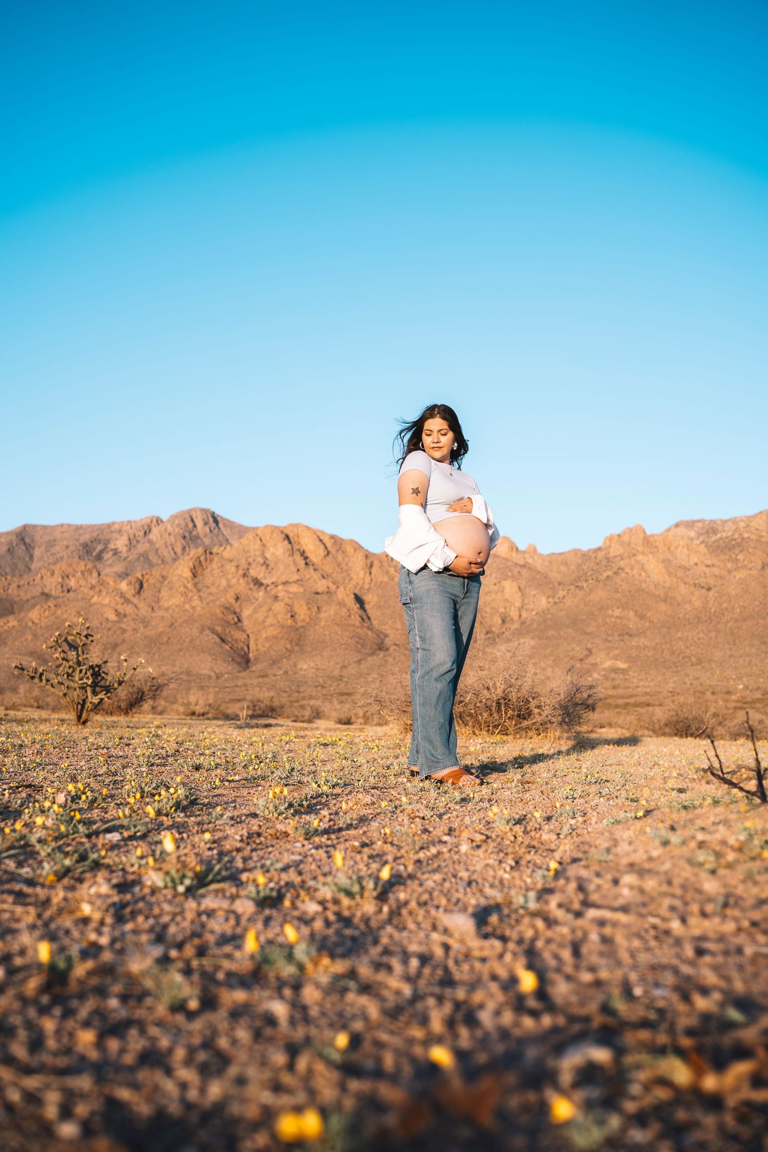 Smiling pregnant woman cradling belly in vast desert landscape with mountains and clear blue sky.