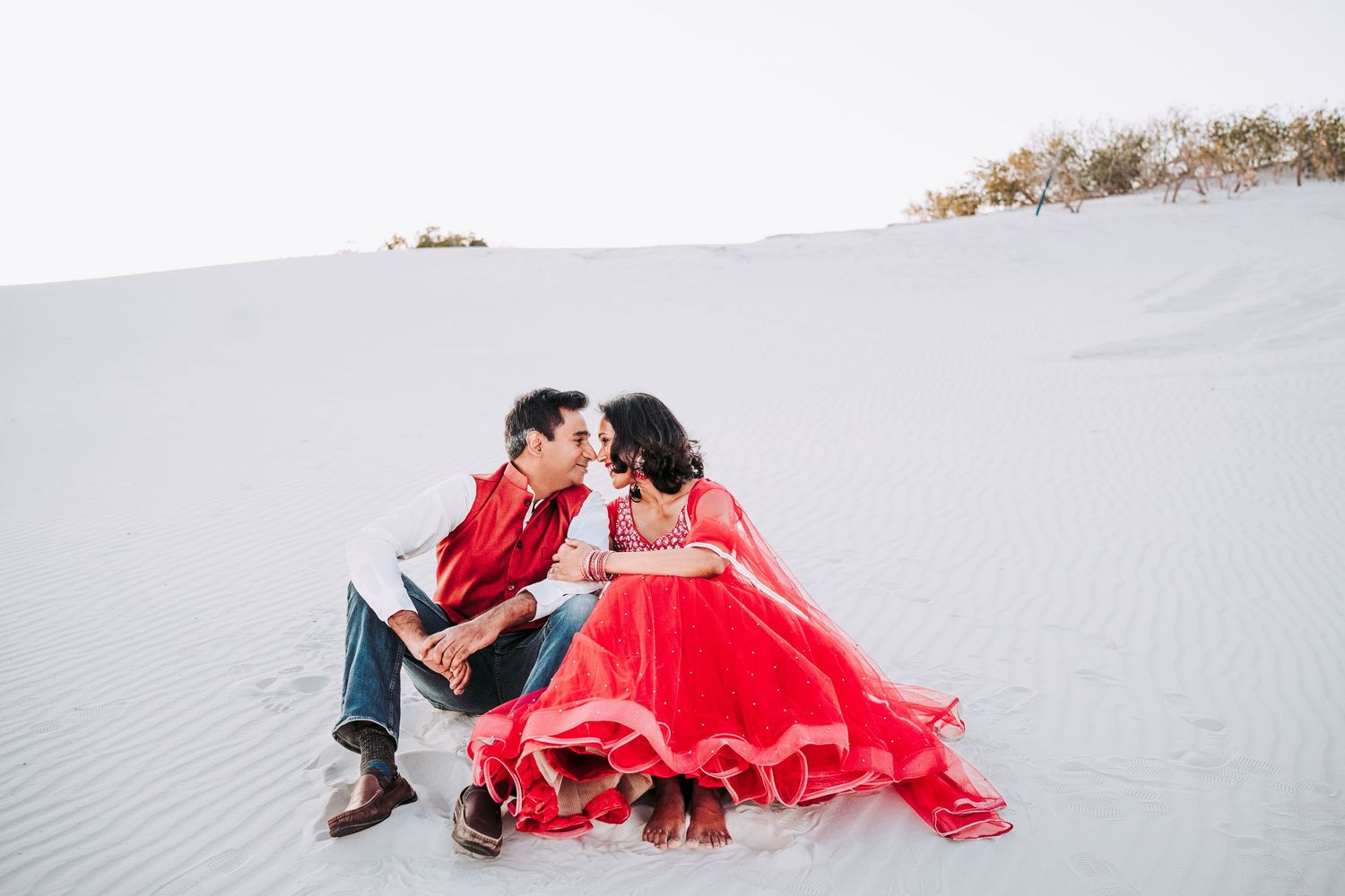 Indian couple in red traditional outfits sharing a romantic moment on white sand dunes.