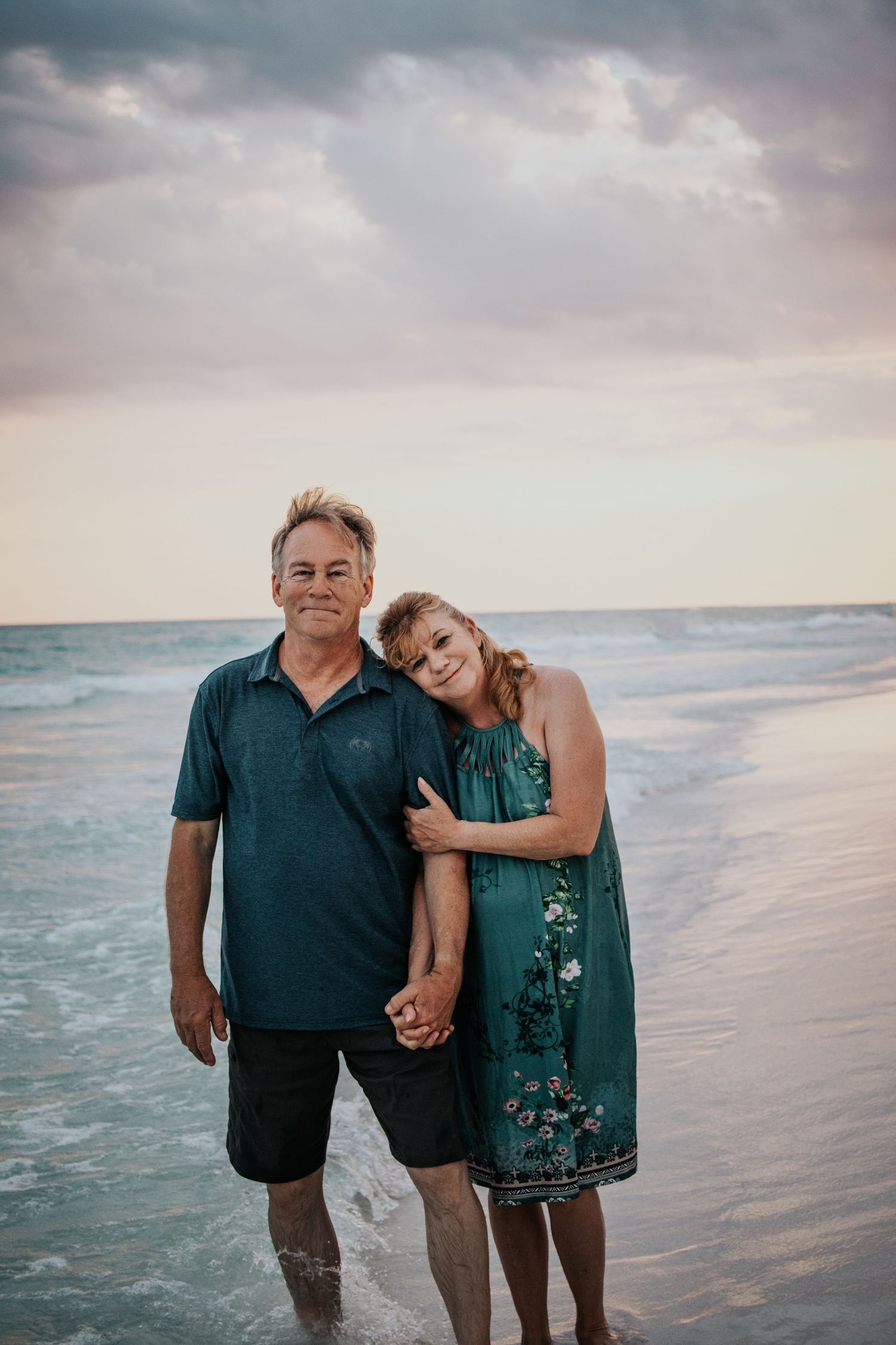 Smiling couple holding hands and posing together on a beach with turquoise ocean waves behind.