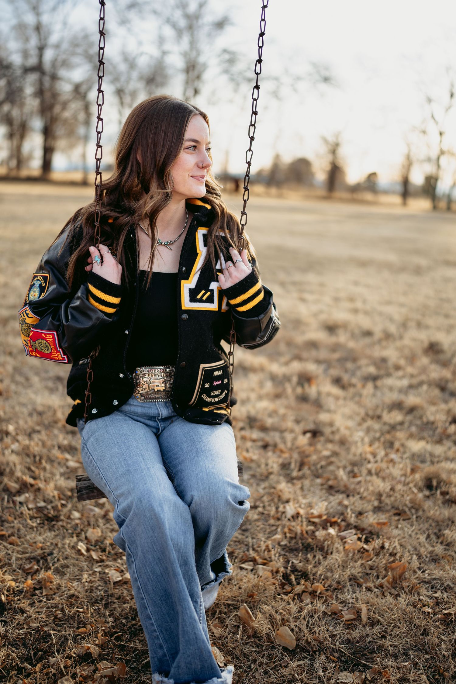 High school senior girl sitting on swing wearing black and gold letterman jacket in autumn field.