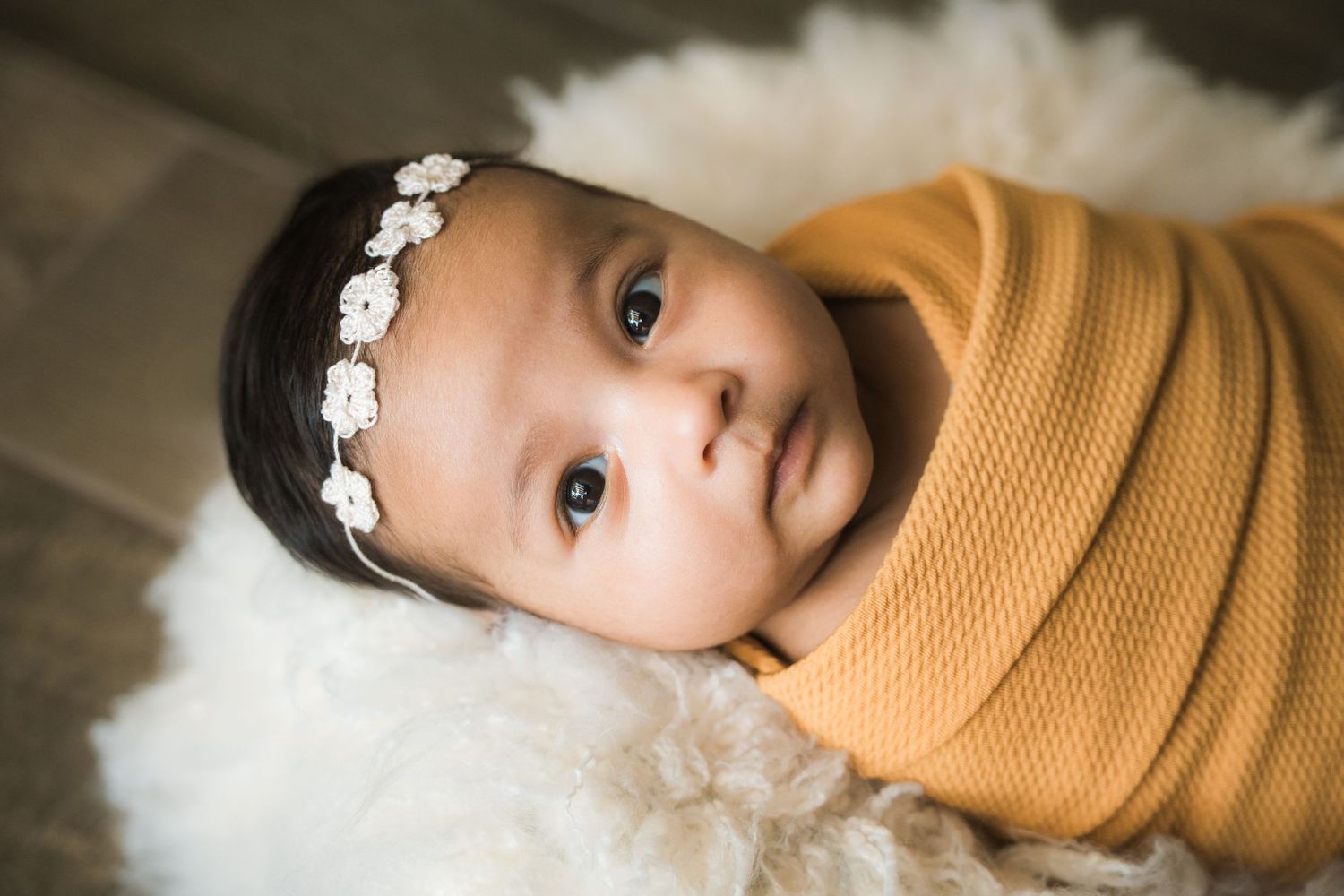 Close-up portrait of wide-eyed newborn baby girl in mustard swaddle with floral headband.