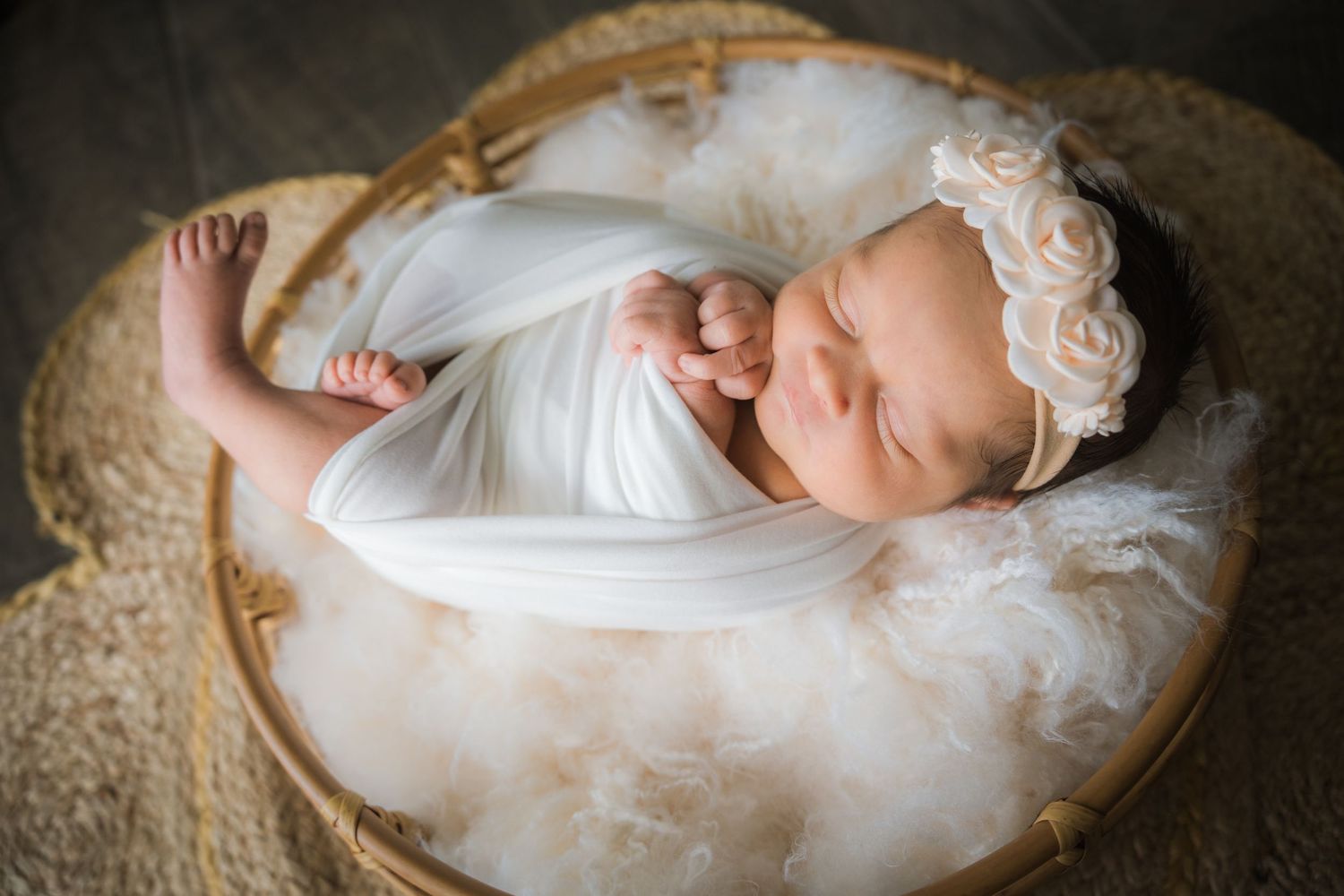 Newborn baby girl swaddled in white wrap with rose headband sleeping in wooden bowl with white feathers.
