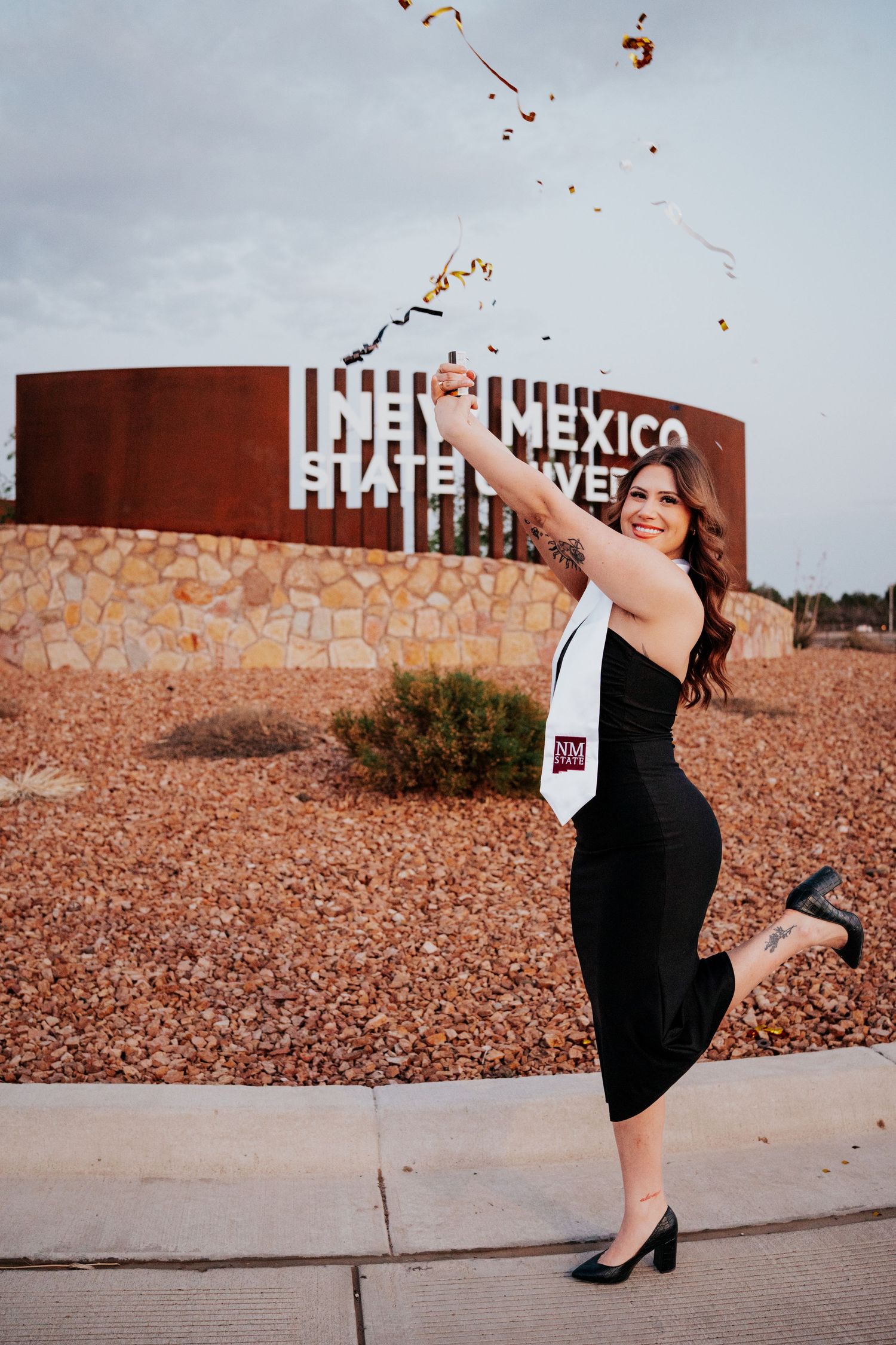 NMSU graduate celebrating in black dress with confetti by New Mexico State University sign.