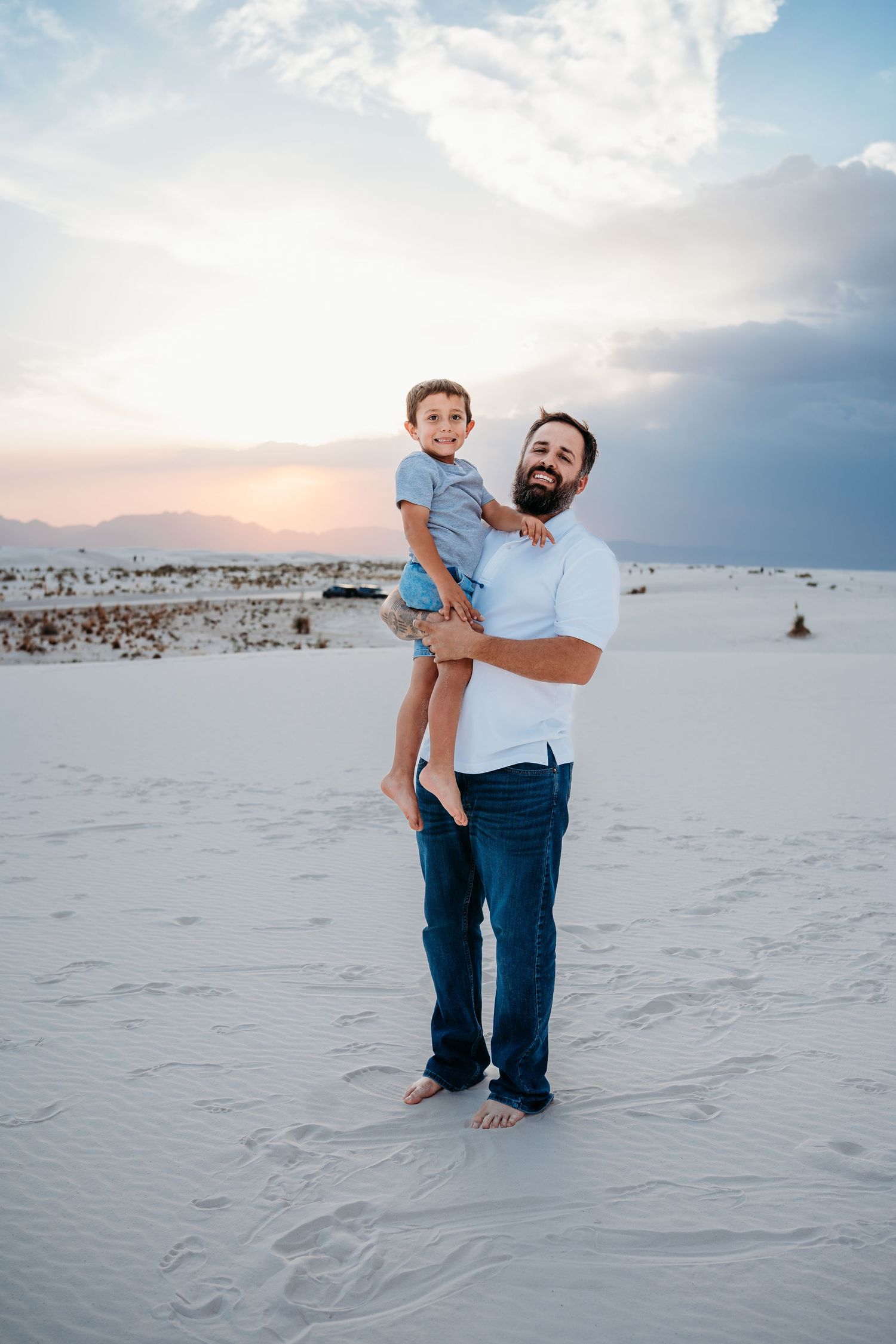 A bearded father smiles proudly while holding his young son on white sand dunes at sunset.