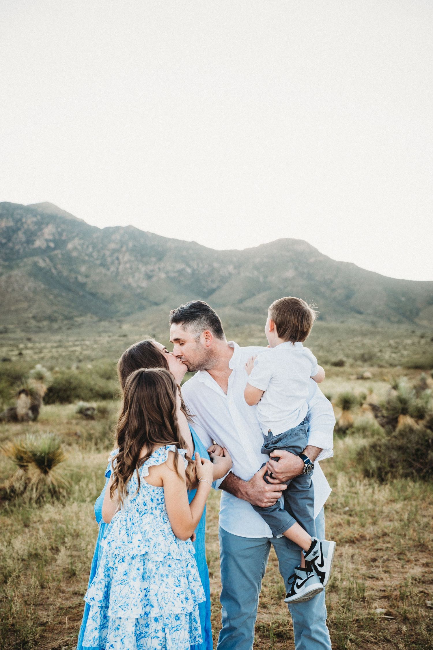 Family of four with parents kissing while dad holds toddler in desert.