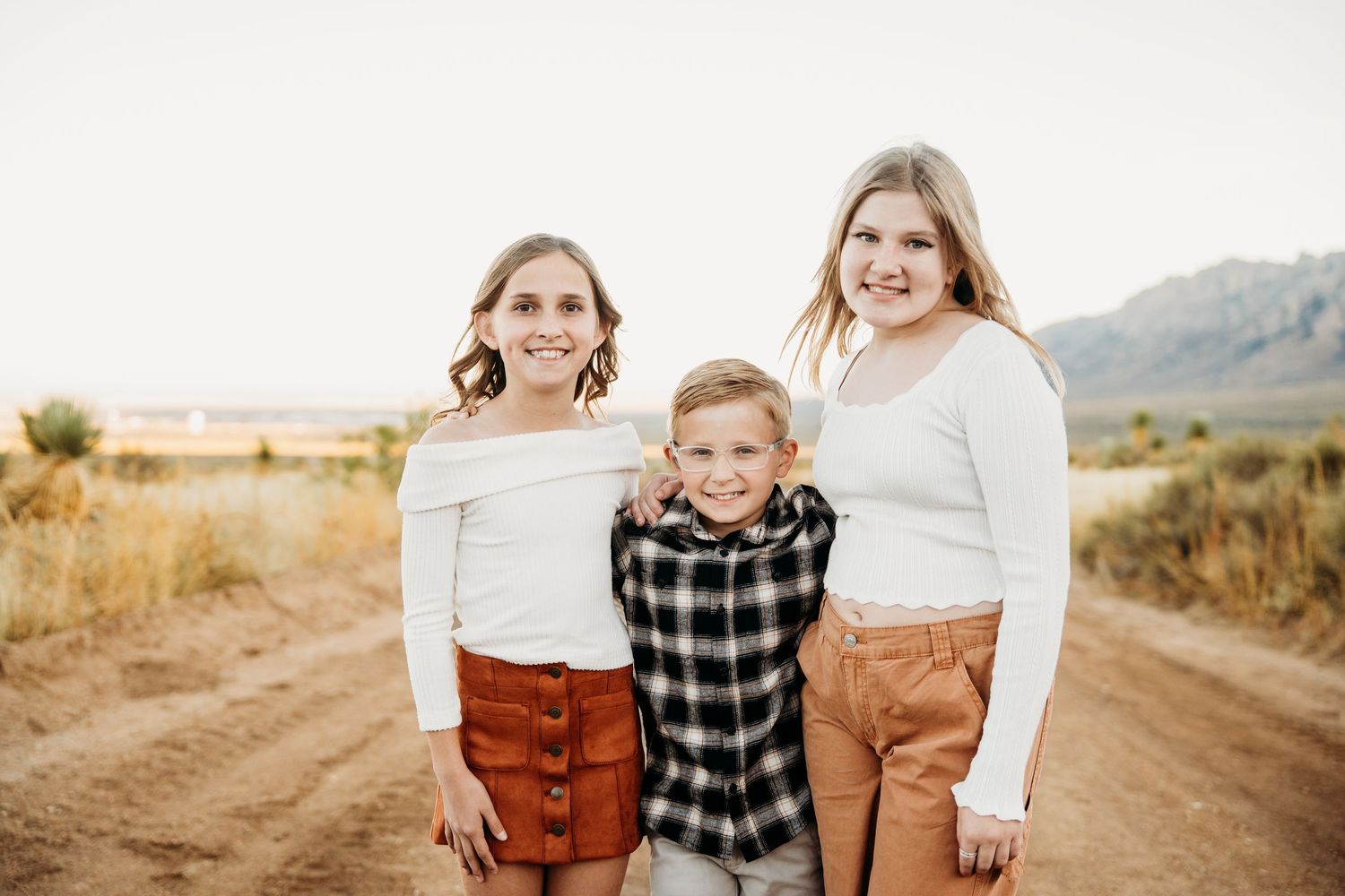 Three children pose together smiling on a dirt road with mountains in the background.