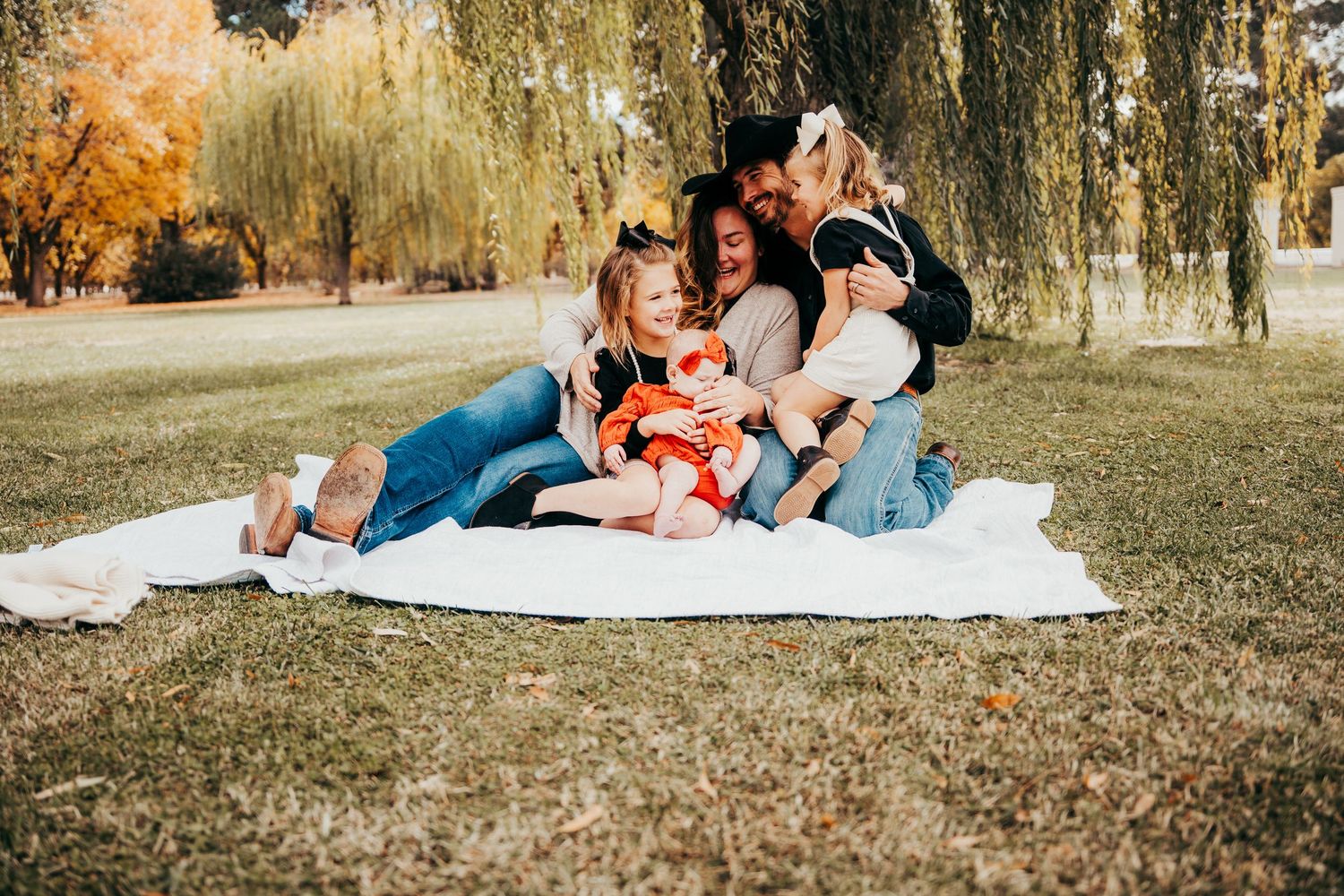 Family of four snuggling on white blanket under willow tree during autumn family photo session.