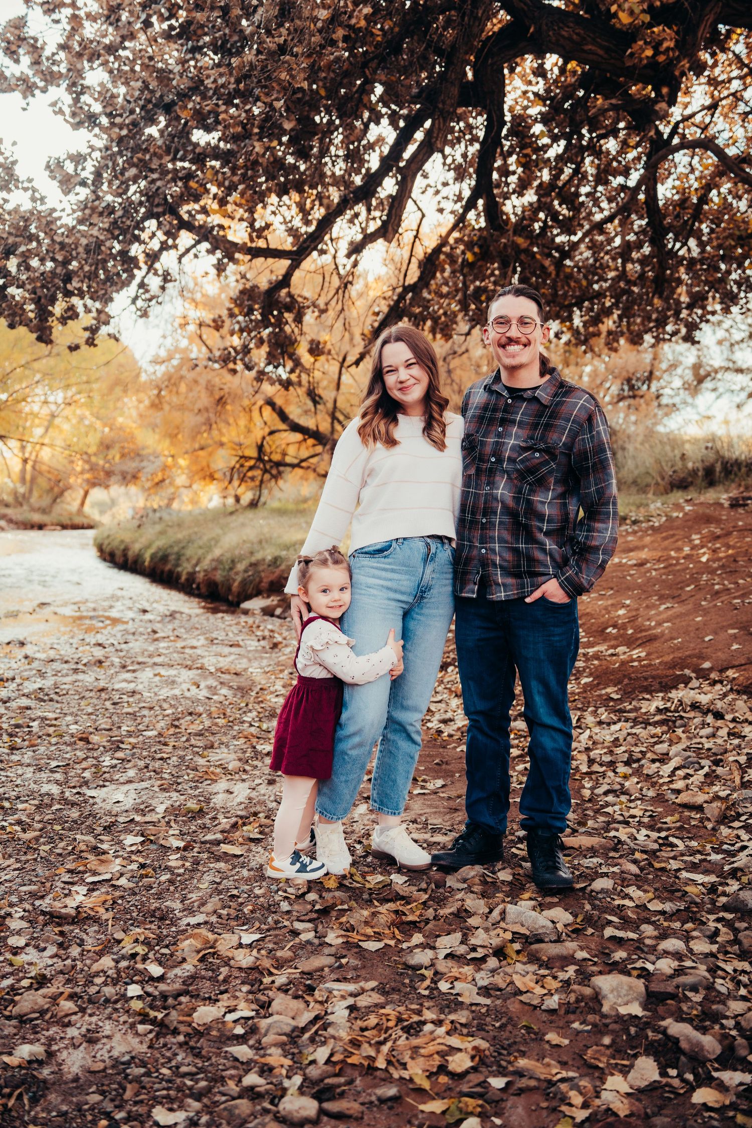 Family of three posing outdoors among fallen autumn leaves with golden trees and creek behind them.