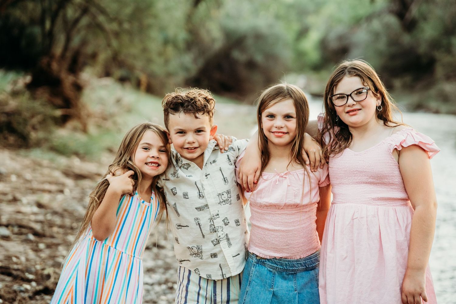 Four siblings smile together in a casual outdoor portrait by a creek.
