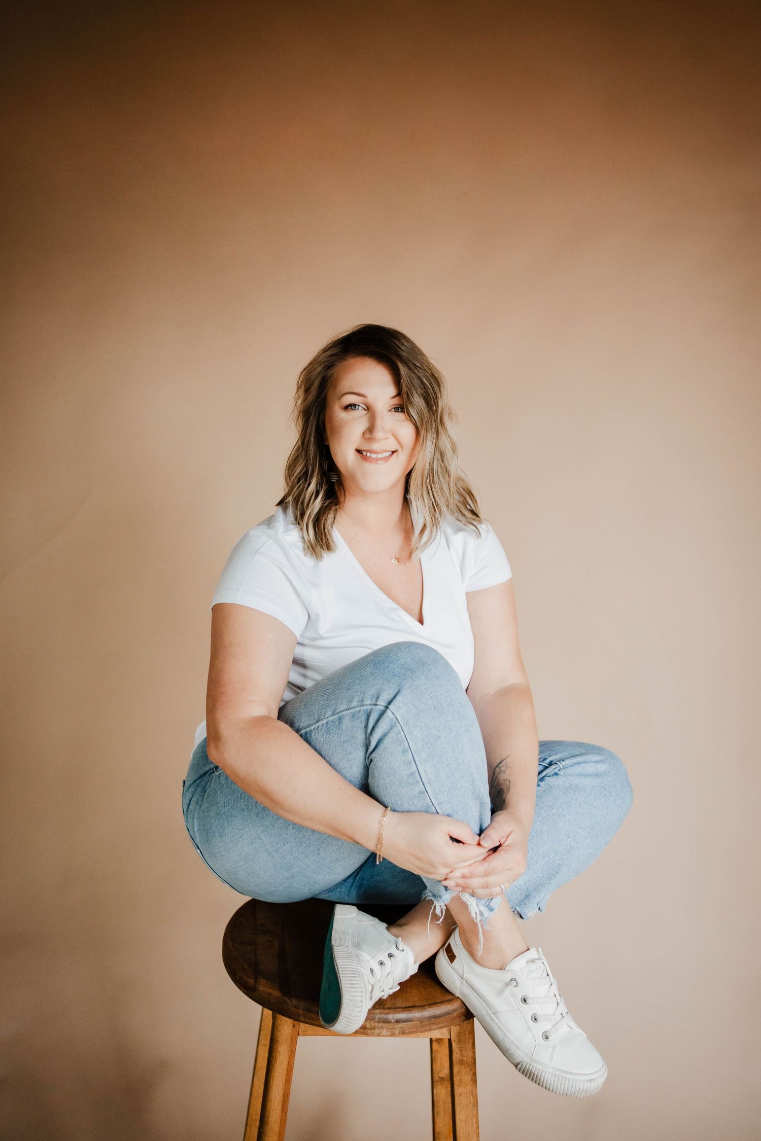 Woman in white t-shirt and jeans sitting cross-legged on a wooden stool against a warm beige background.
