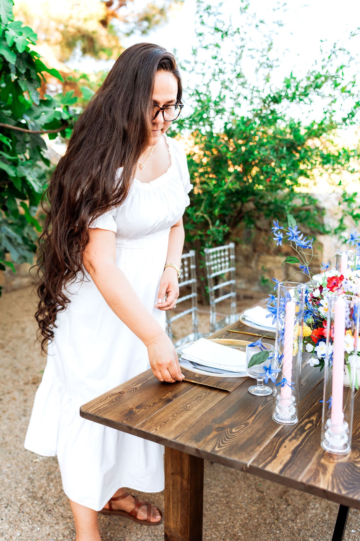 Brunette woman in white dress arranging place settings at an outdoor wedding reception table with blue floral decor.