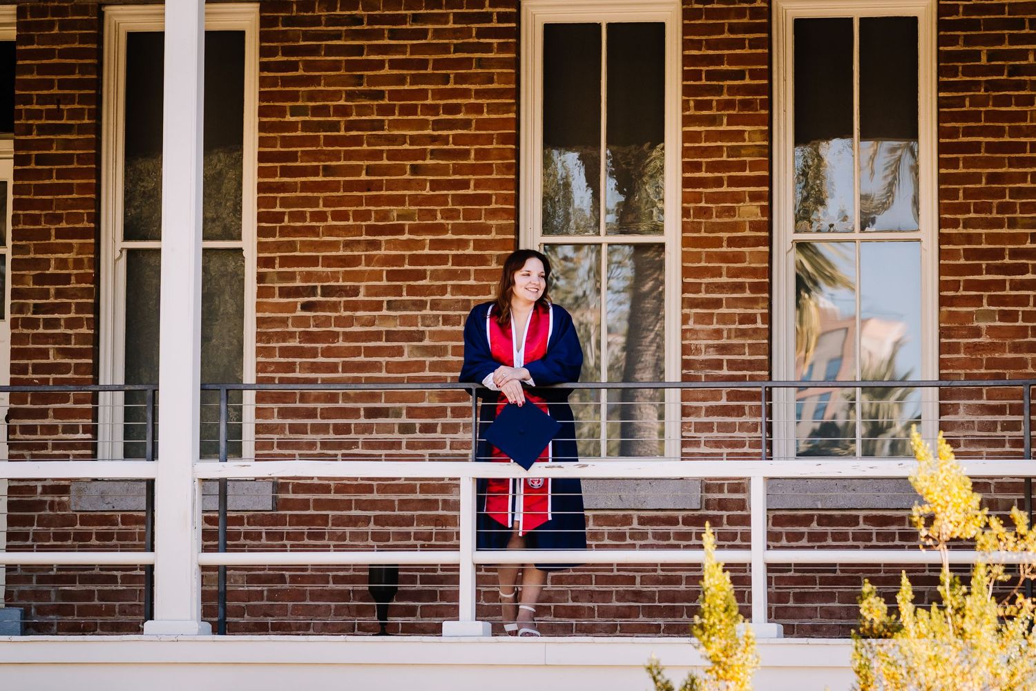 Woman in navy graduation gown stands on brick building porch, holding red stole, with tree reflections in windows behind her.