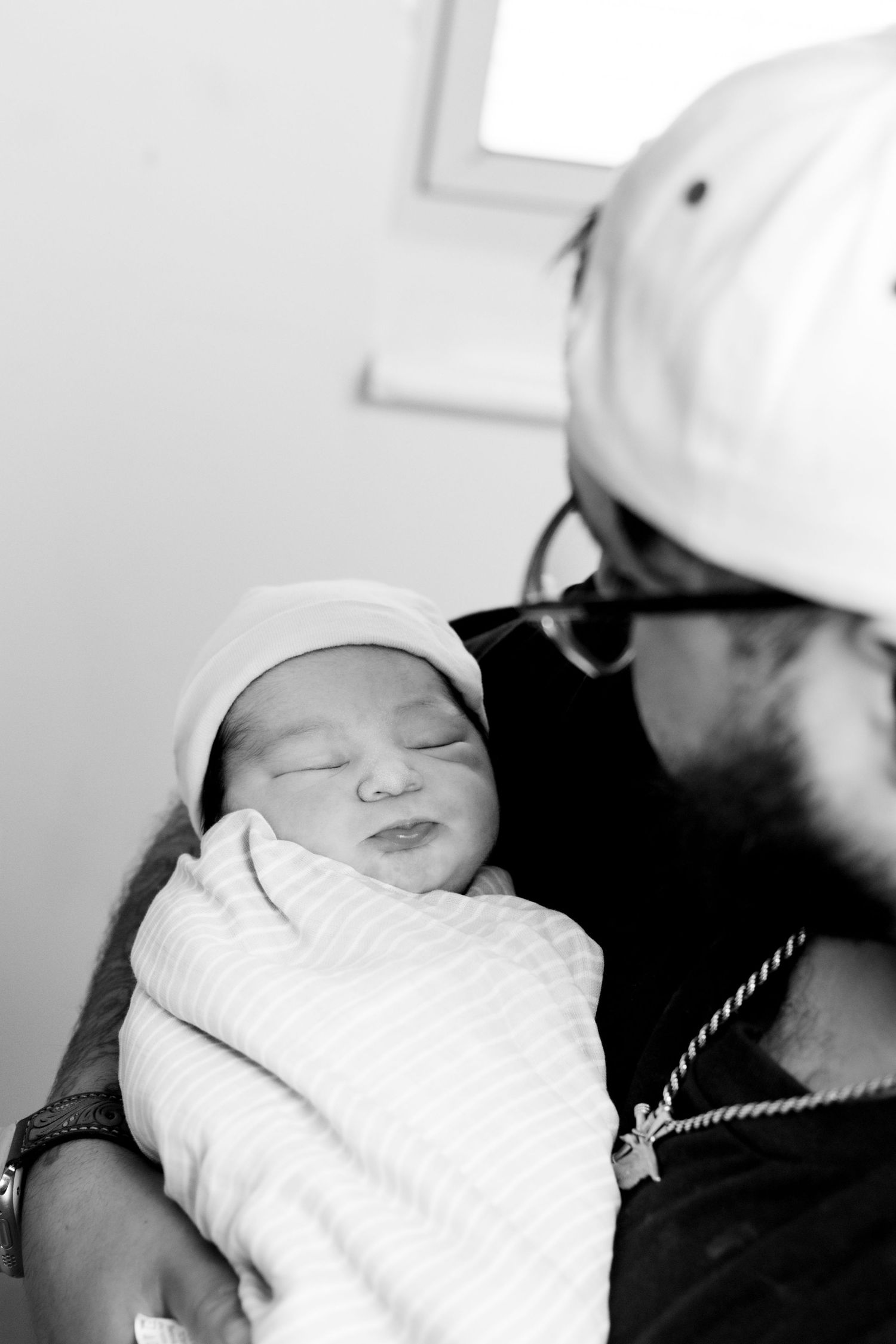 Black and white image of a parent in a cap holding a peacefully sleeping swaddled newborn.