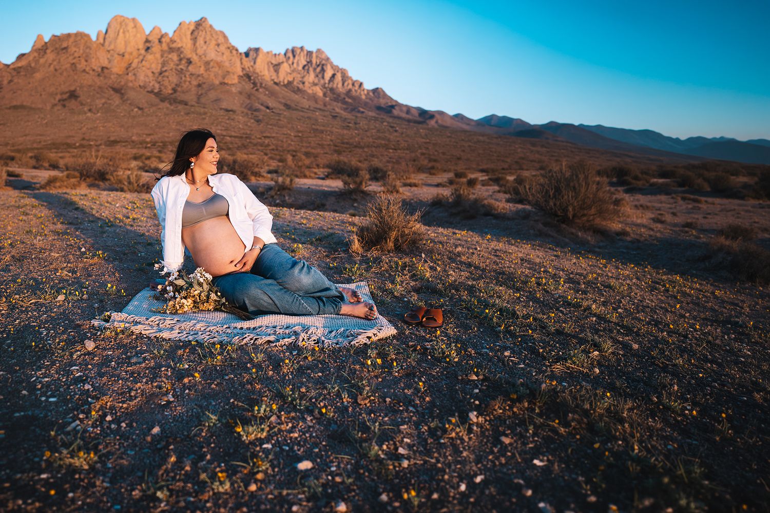 Pregnant woman relaxing on blanket in desert landscape with rugged mountains at golden hour.