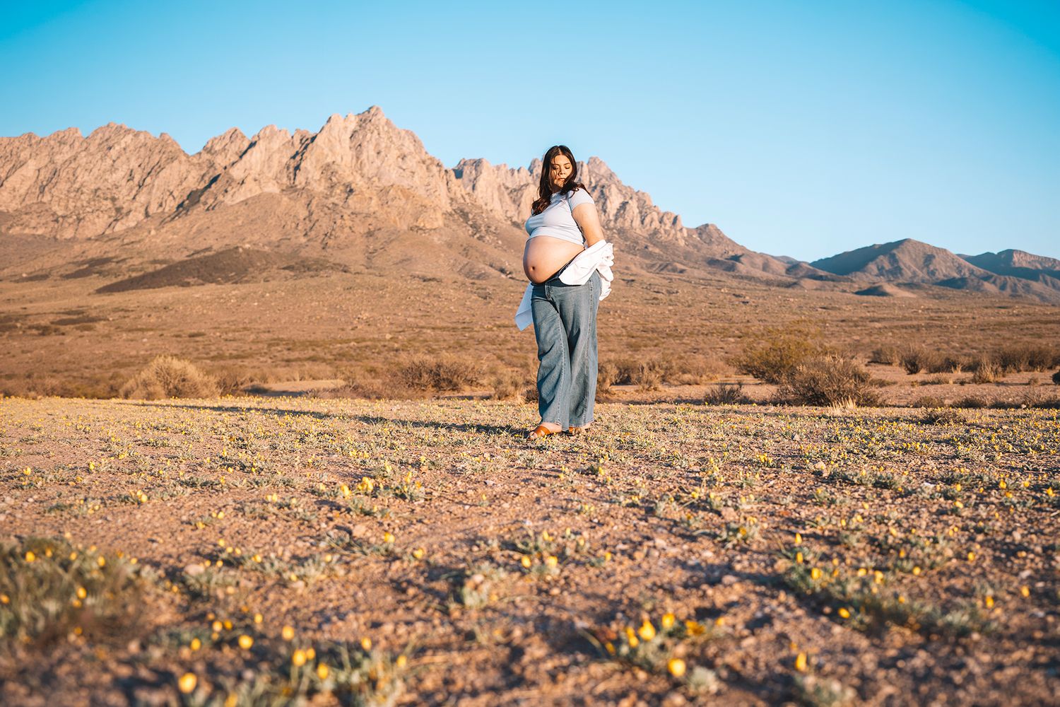 Pregnant woman in white top and wide-leg jeans poses in vast desert field with rocky mountains behind.