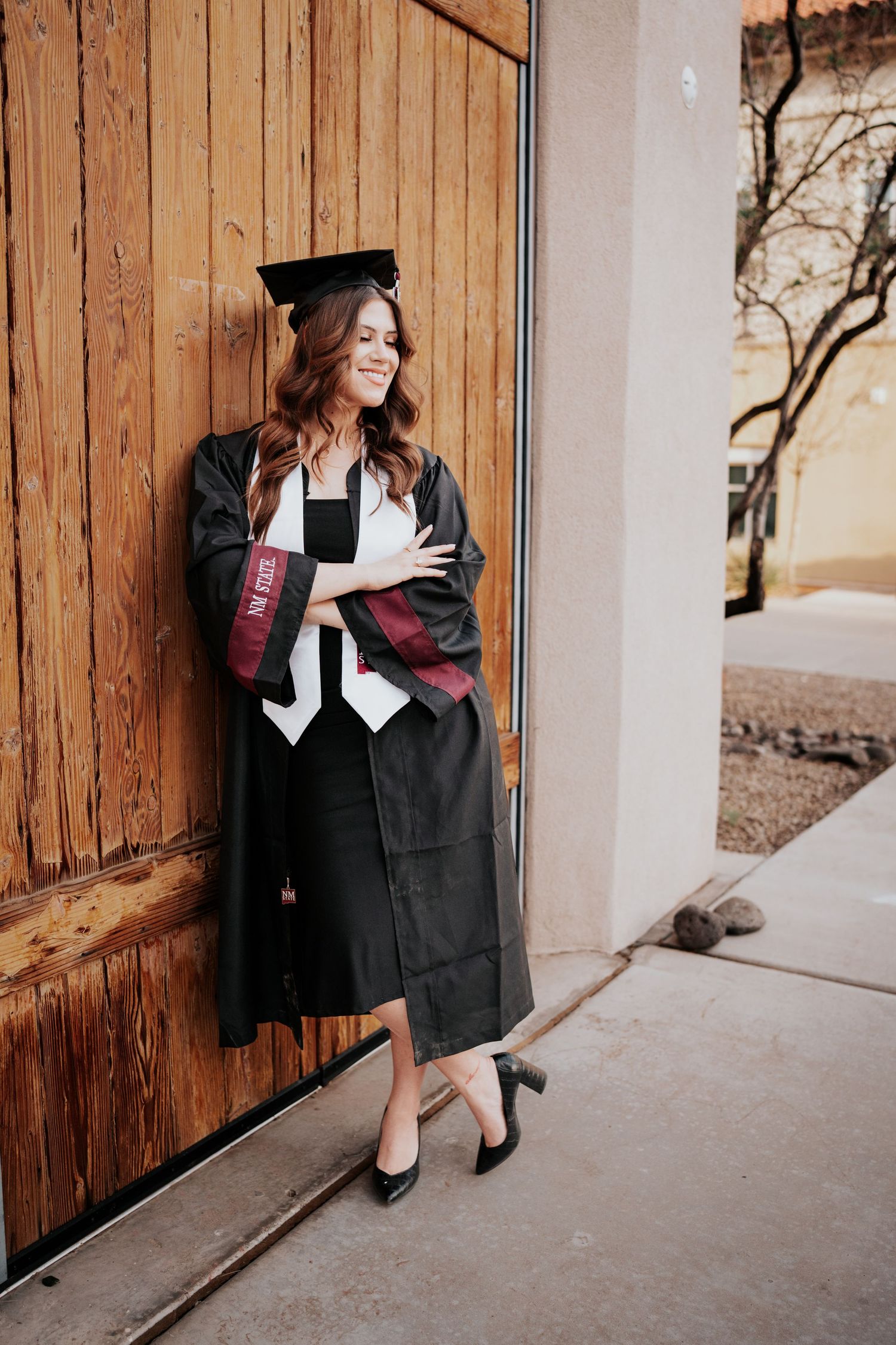 Female graduate in full cap and gown with NM State regalia leaning on wooden door outdoors.