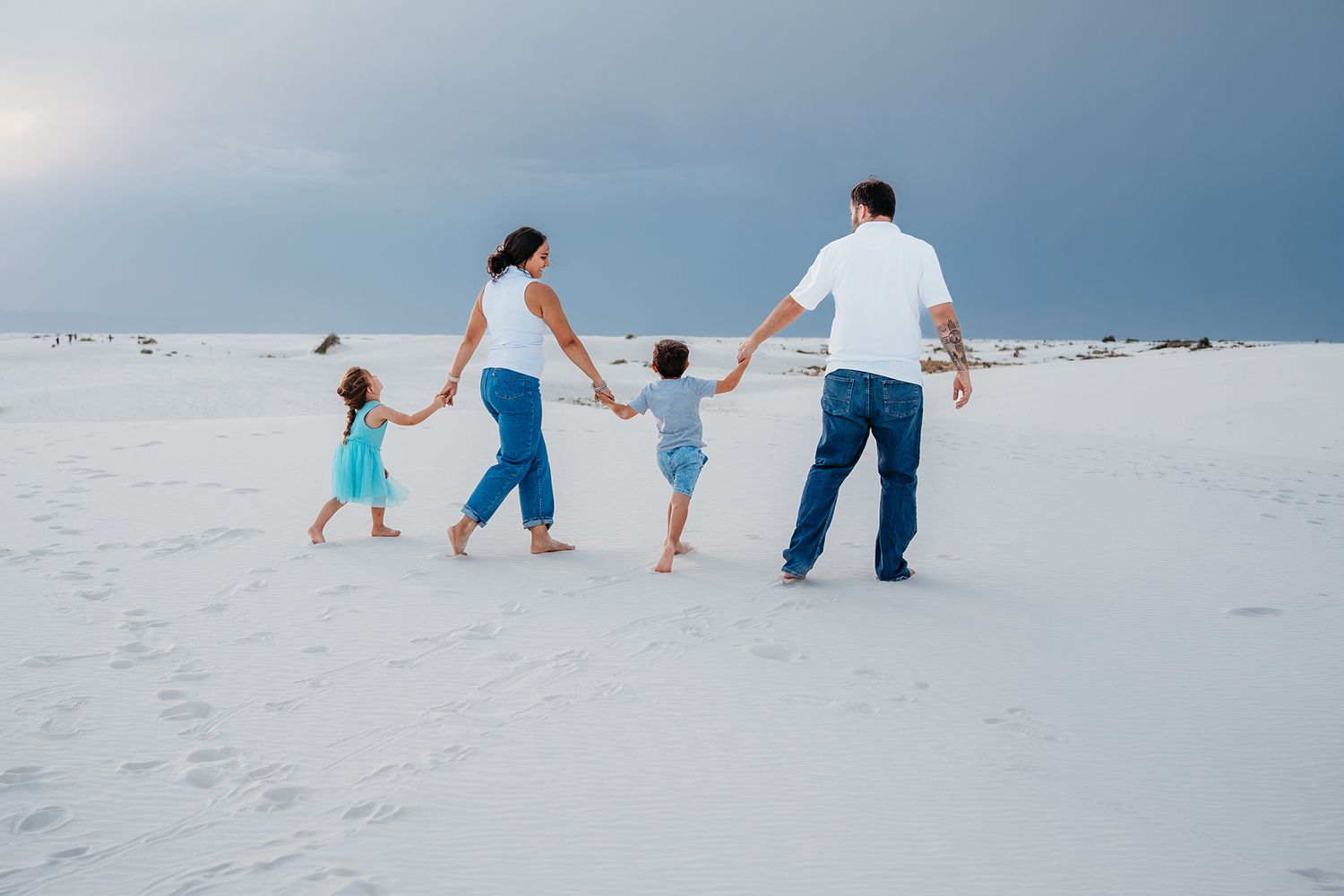 Family of four walks together holding hands across bright white sand dunes under stormy skies.