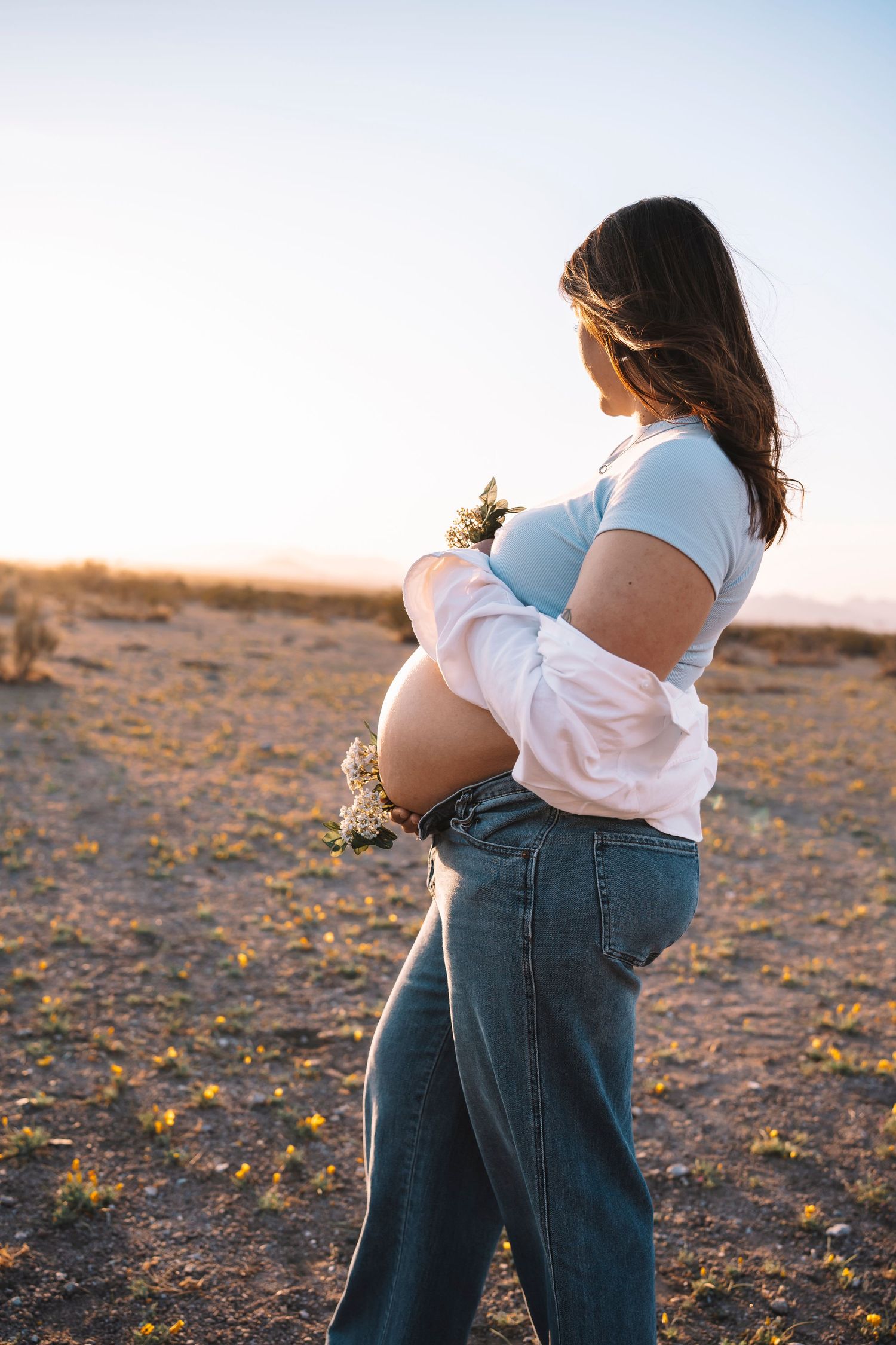 Pregnant woman in white shirt and jeans holding her bare baby bump with wildflowers in a sun-drenched desert landscape.