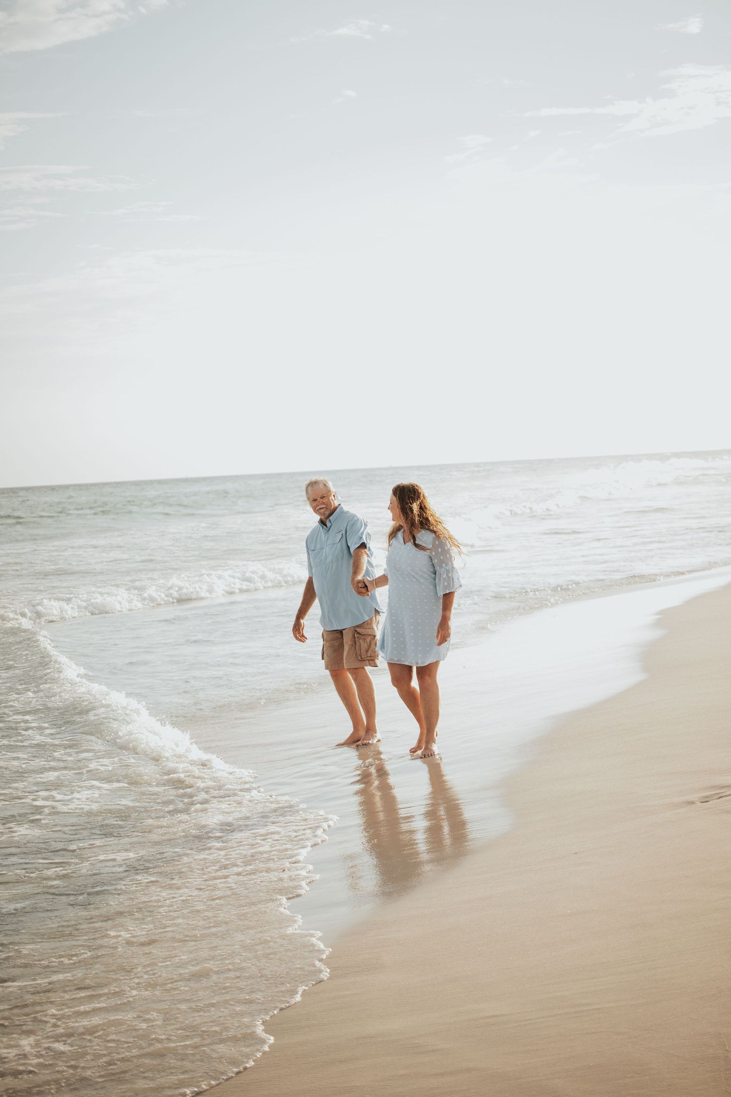 Mature couple holding hands and walking barefoot along a sandy beach with gentle ocean waves nearby.