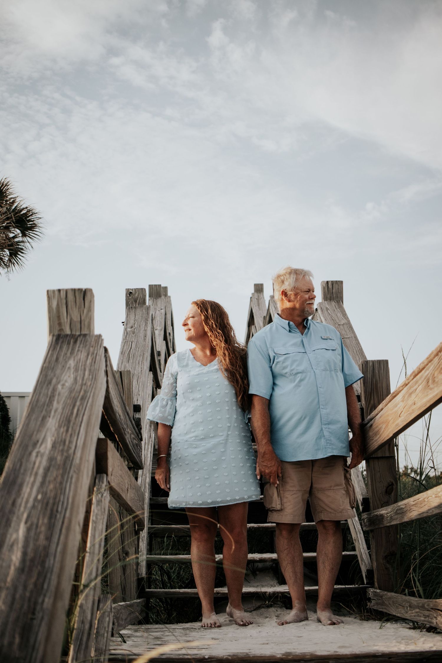 Couple standing hand in hand on wooden beach boardwalk stairs, smiling and looking sideways at sunset.