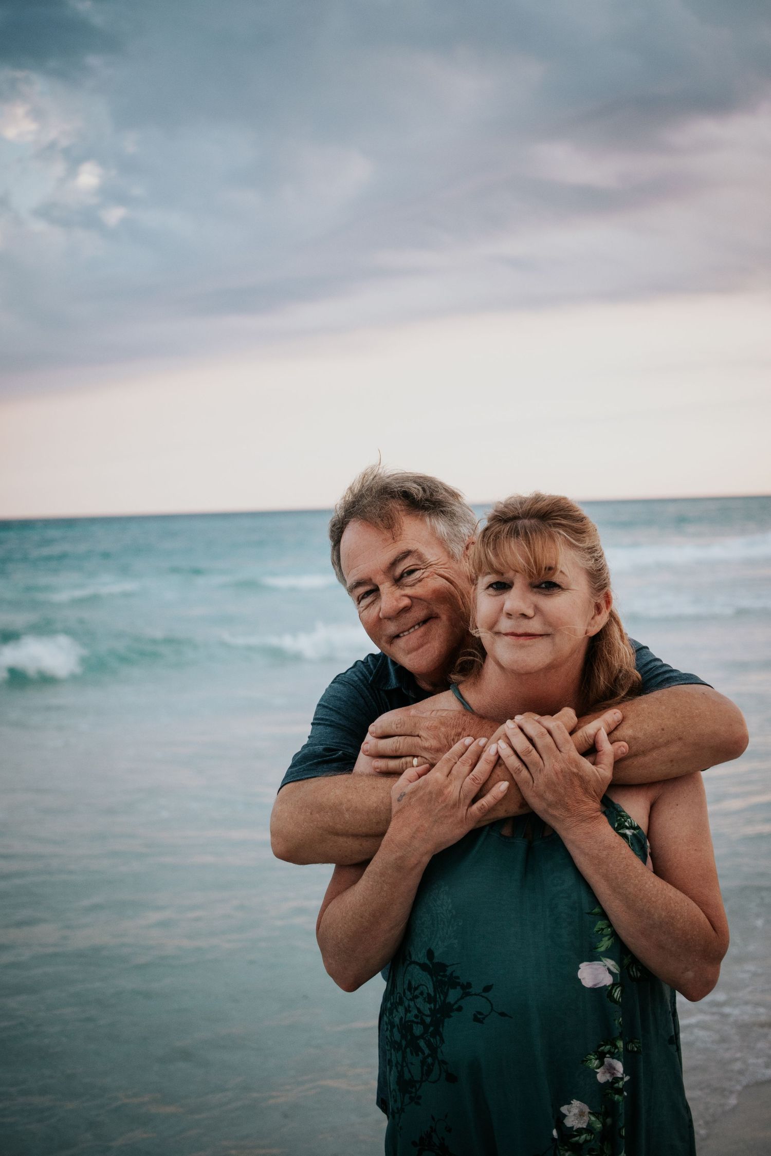 Man hugging woman from behind as they smile at the camera on a beach at dusk.