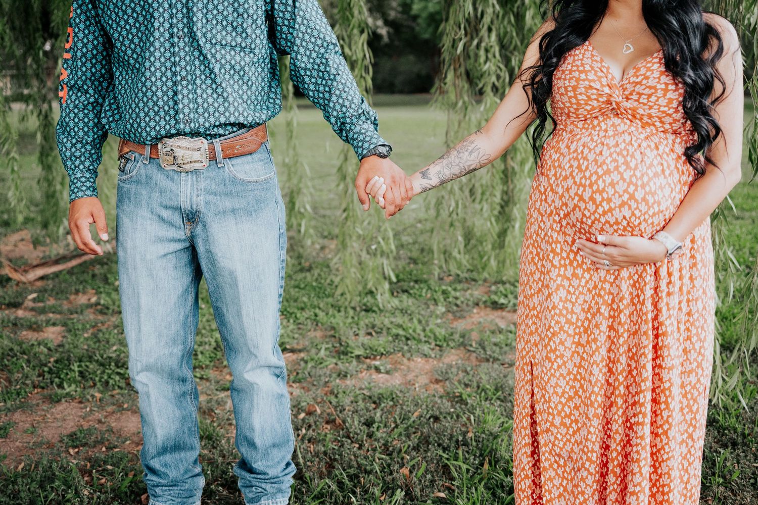 Pregnant woman in orange floral dress holding hands with man in teal shirt and jeans with western belt buckle.