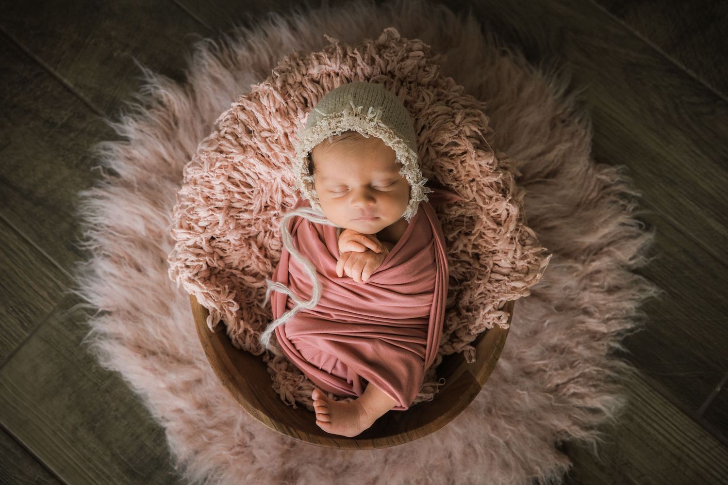 Newborn baby girl swaddled in pink wrap with floral headband, sleeping in wooden bowl with fluffy pink fur.