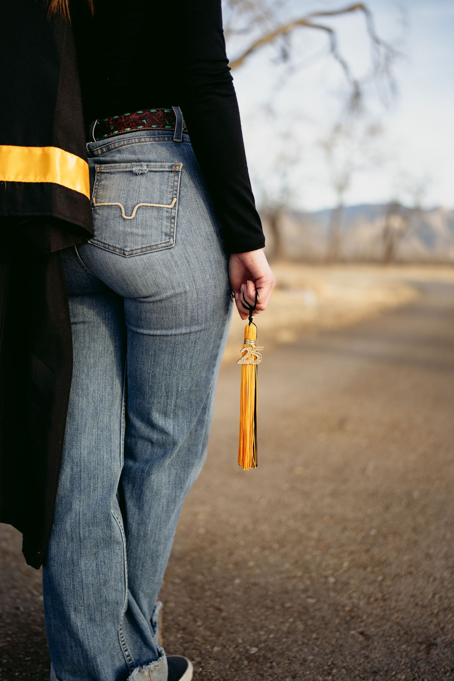 High school senior holding gold graduation tassel wearing jeans and black top with gold accent outdoors.