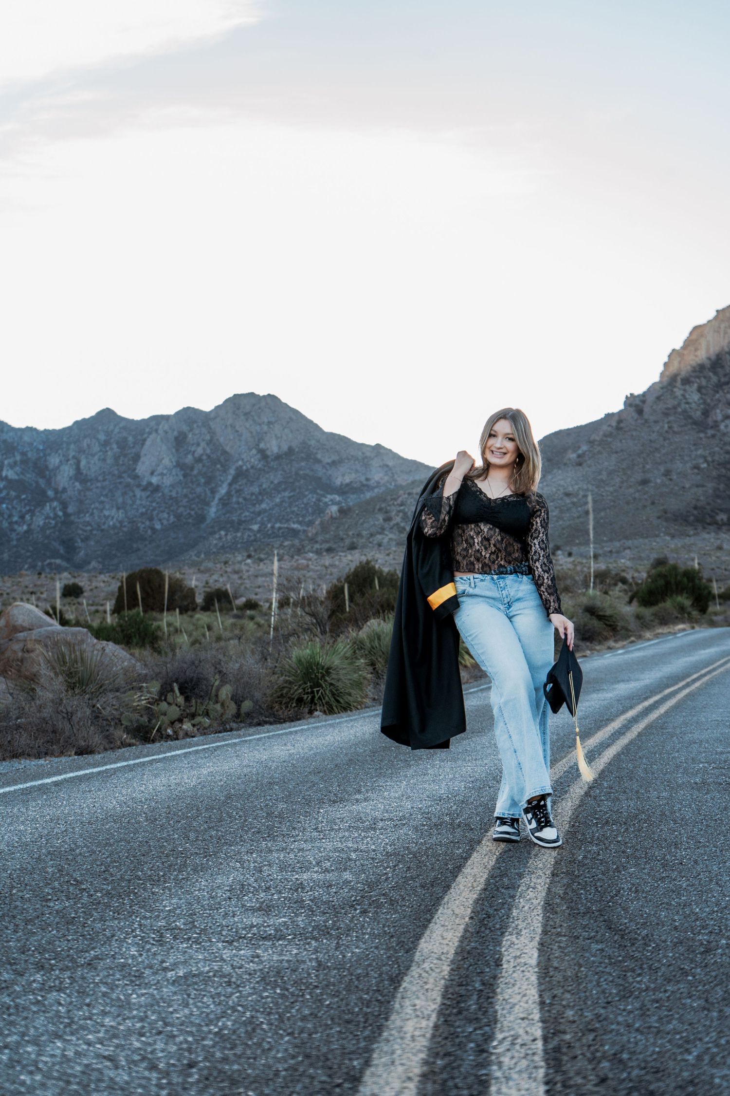 Smiling graduate walks down open road carrying cap while wearing gown over lace top and jeans.