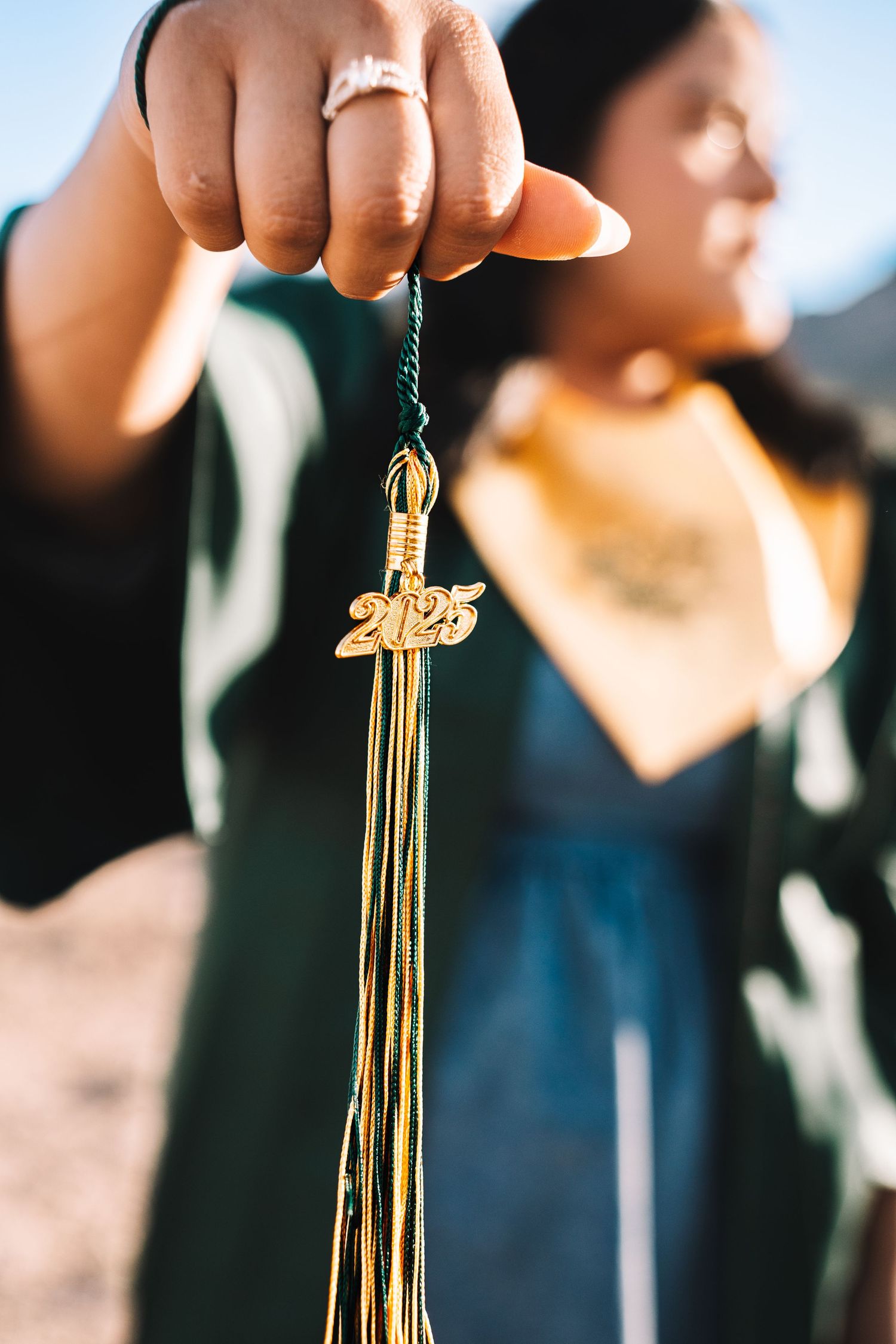 Close-up of a 2025 gold graduation tassel held by a graduate in a green cap and gown outdoors.