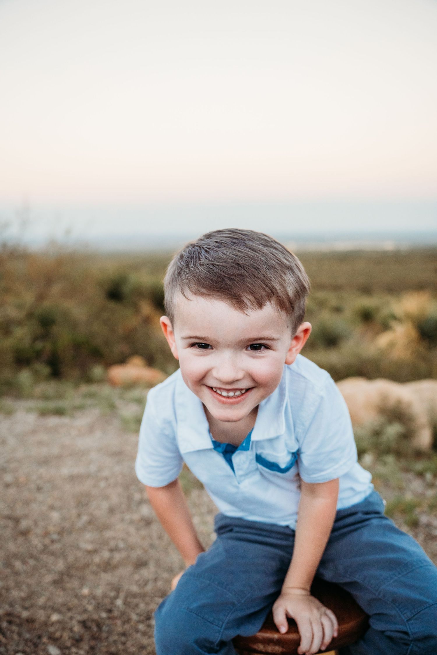 Happy young boy leaning forward and smiling outdoors with an open sky and field behind him.