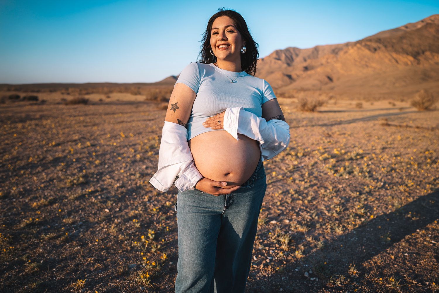 Smiling pregnant woman holds baby bump outdoors in desert landscape at golden hour.