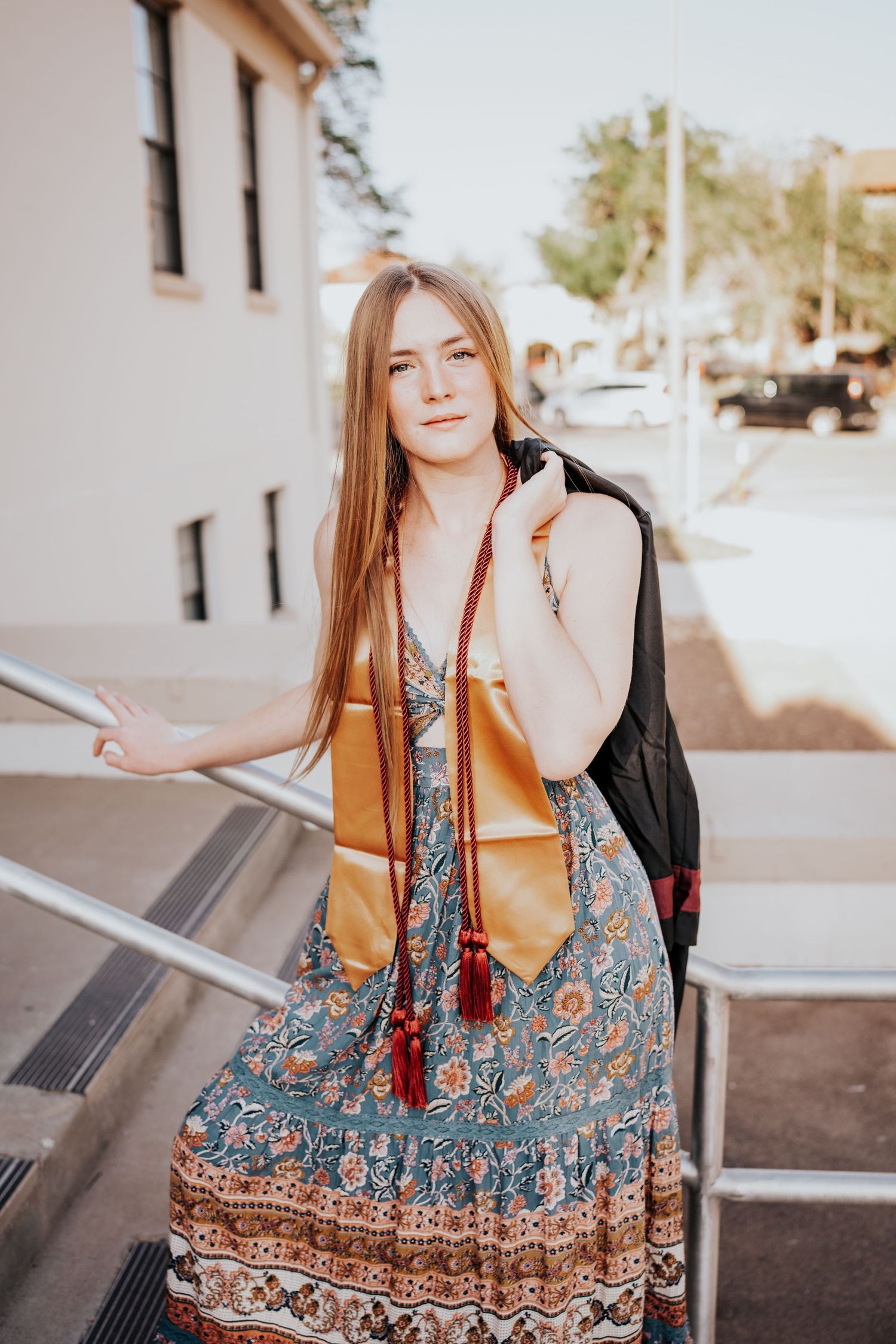 Young woman in floral maxi dress with graduation stole posing on university steps.