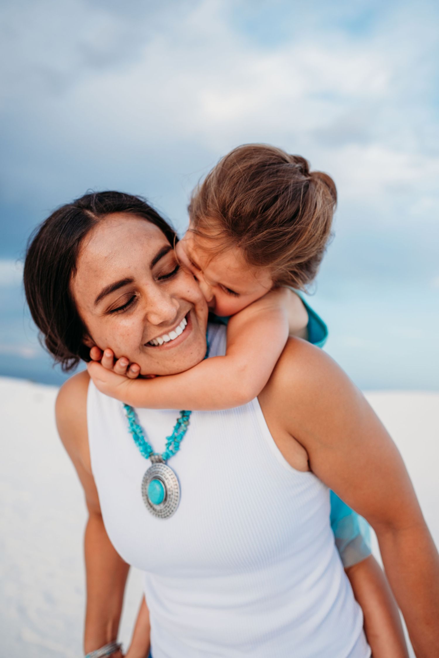 Joyful mother with turquoise pendant necklace receiving kiss from child on white sand beach.