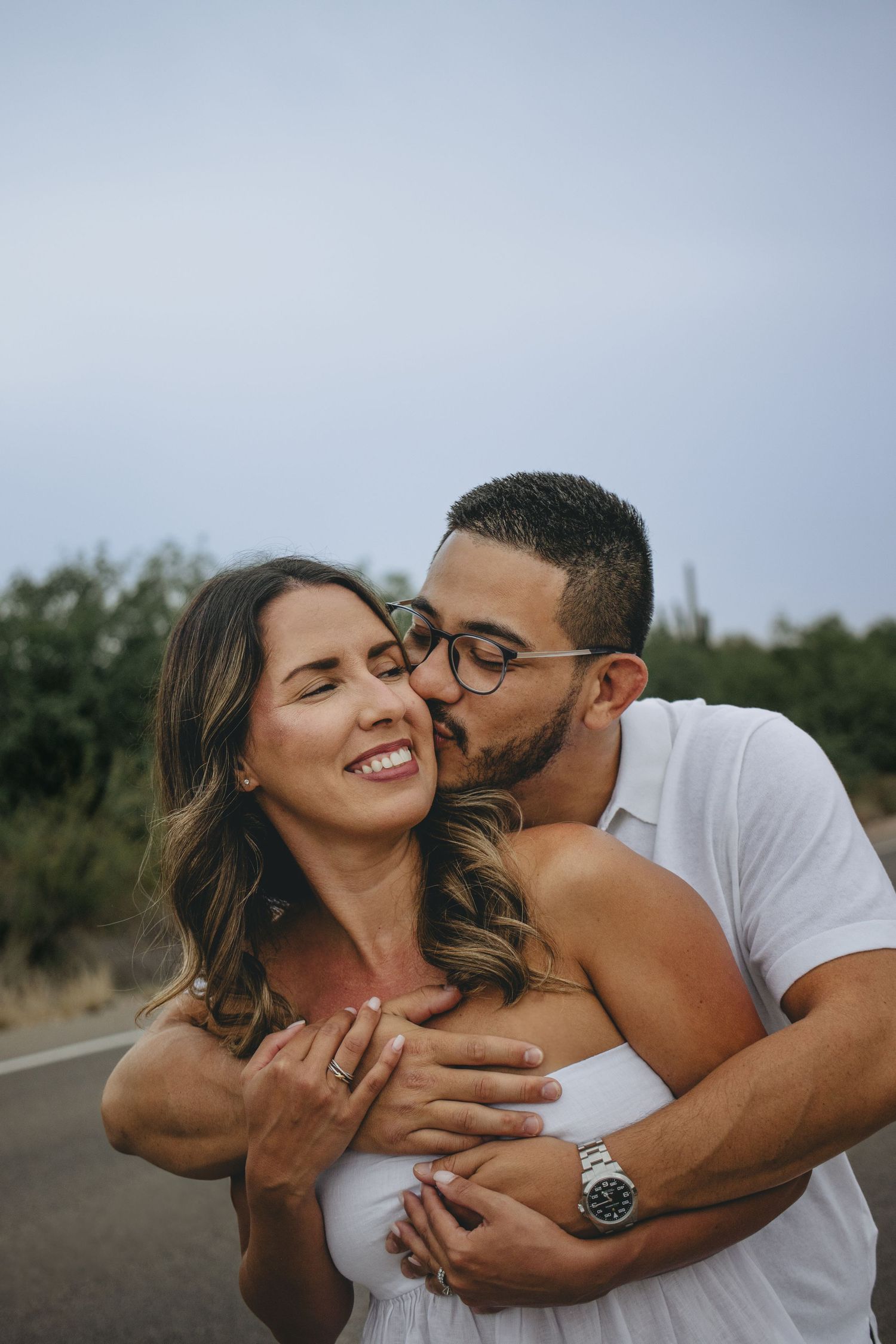 Man kissing smiling woman on cheek while embracing her on a desert road, wearing white outfits.