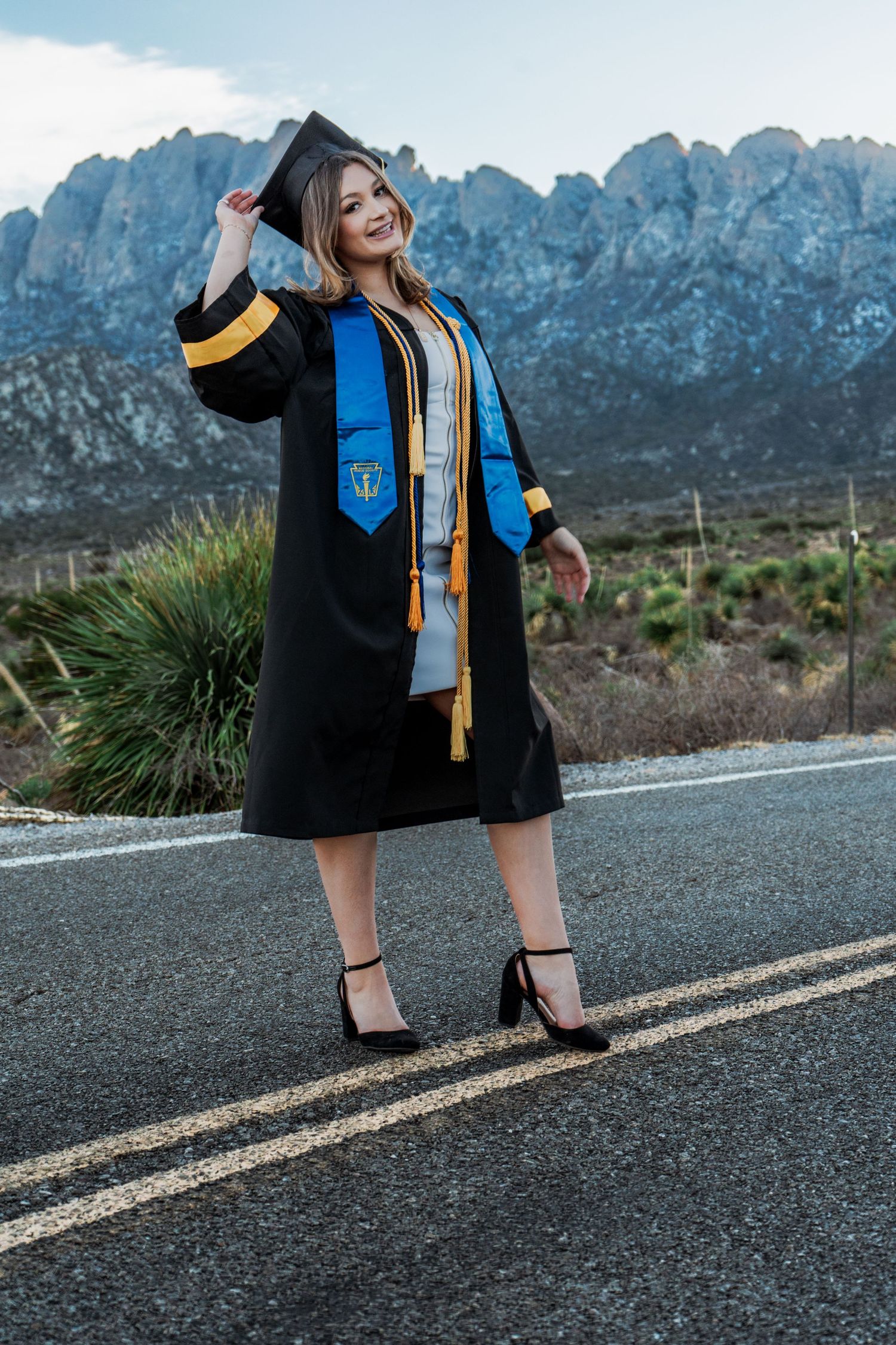Female graduate in black gown with blue honor stole posing on road with dramatic mountain range backdrop.