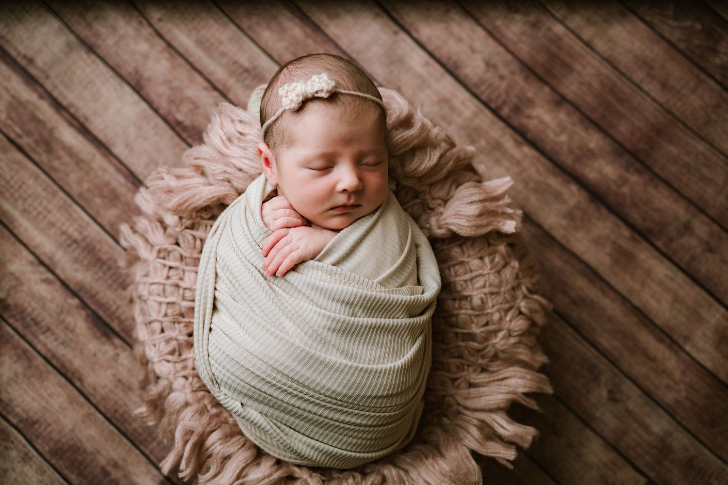 Newborn baby swaddled in green wrap sleeping on fluffy pink fur on wood backdrop.