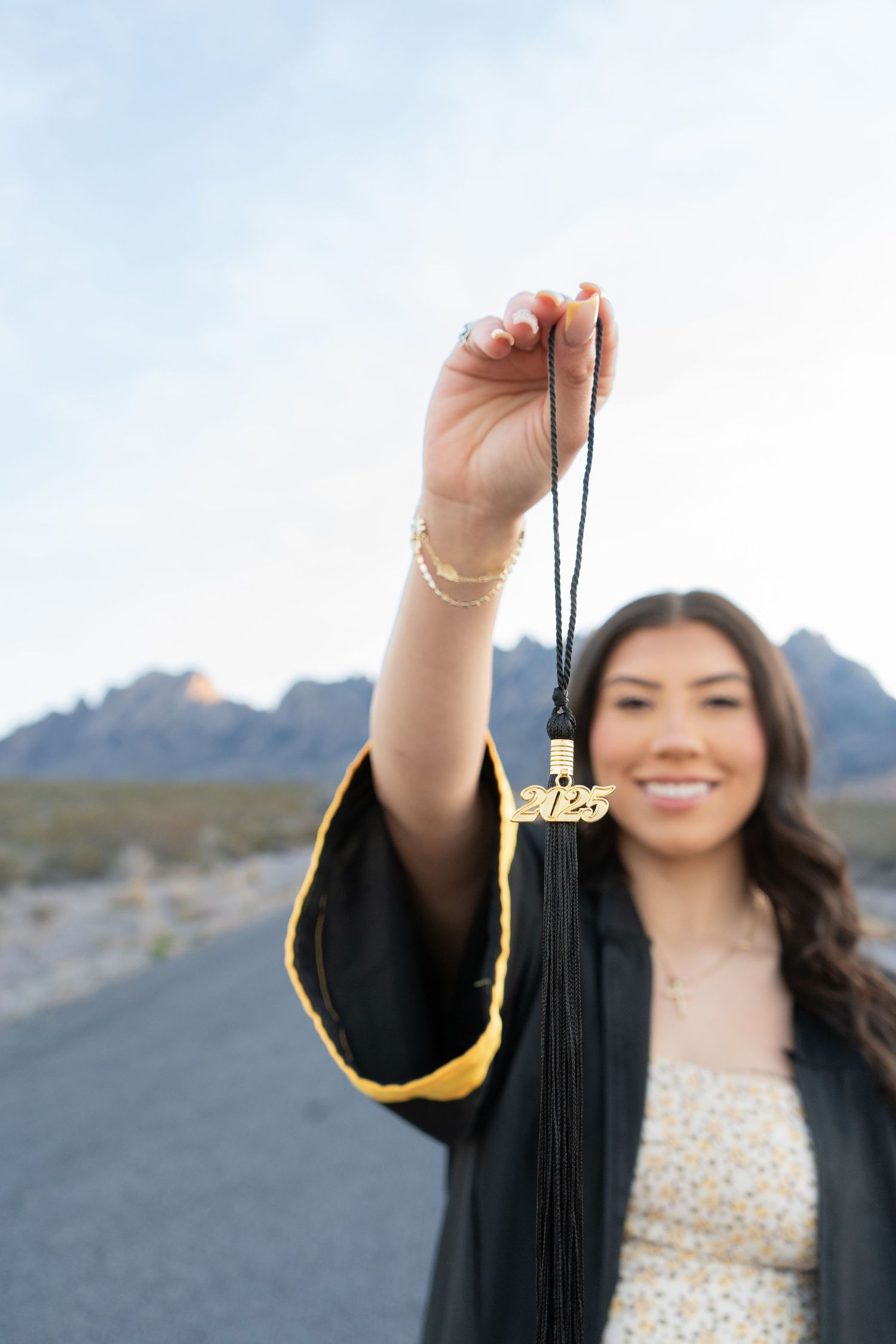 Smiling female graduate in black gown holds up a gold 2025 graduation tassel with mountains in the background.