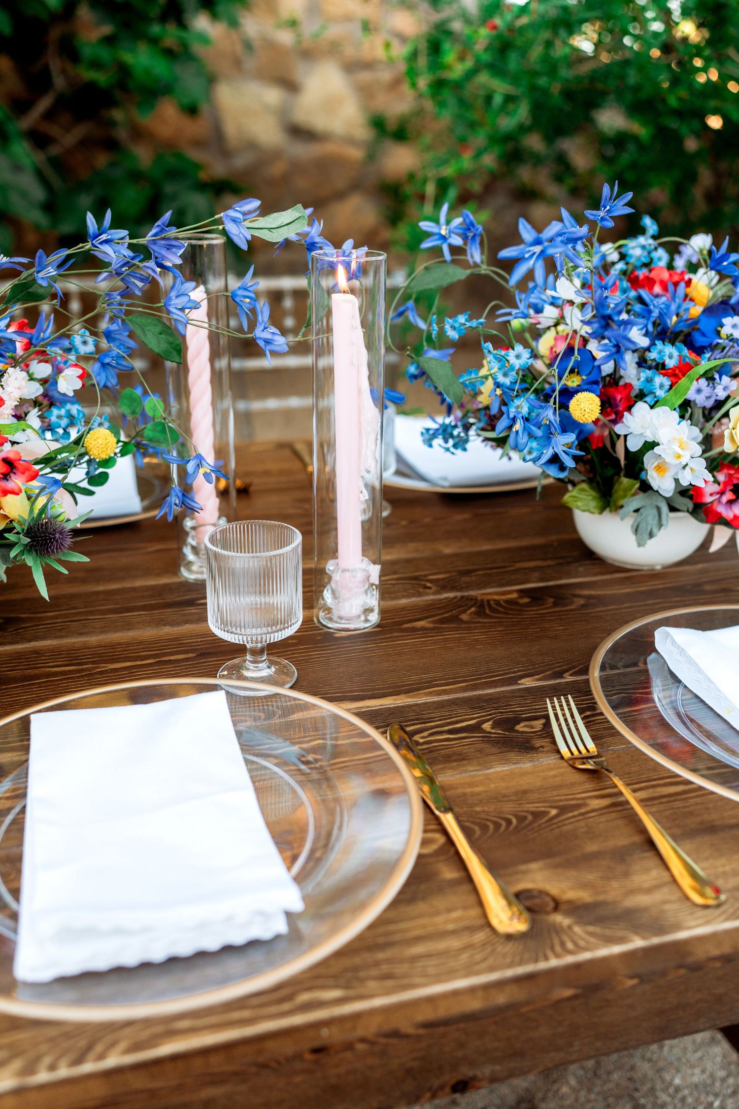 Elegant rustic outdoor place setting with gold flatware, glass charger, and colorful wildflower arrangements.