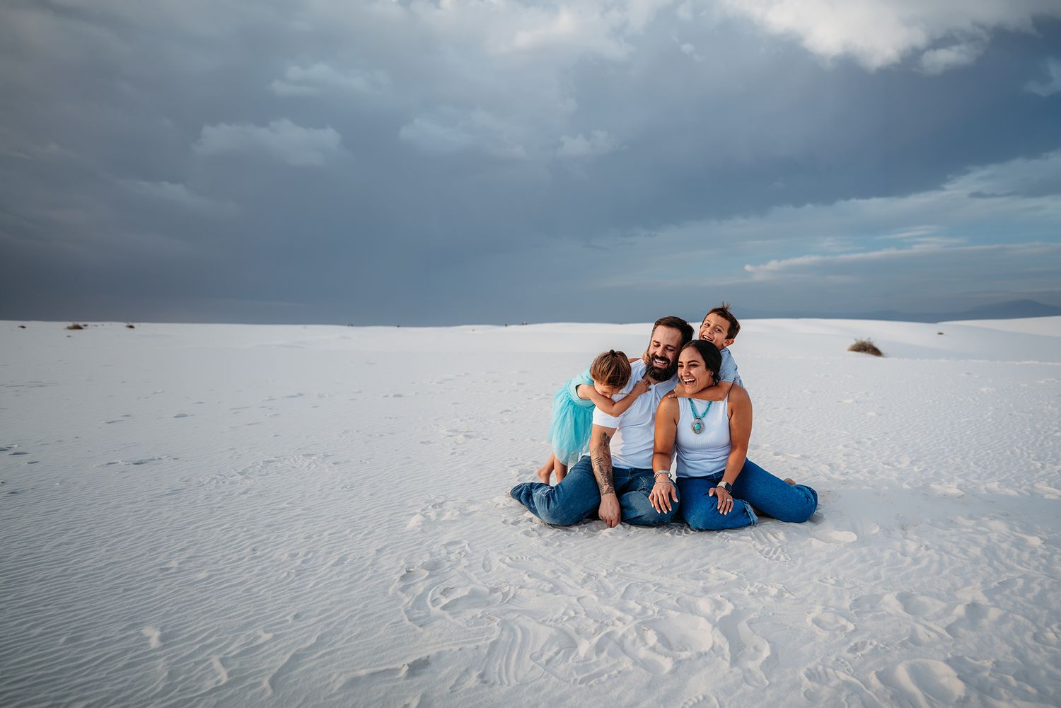 Happy family of four sitting together on white sand dunes, smiling and embracing under cloudy skies.