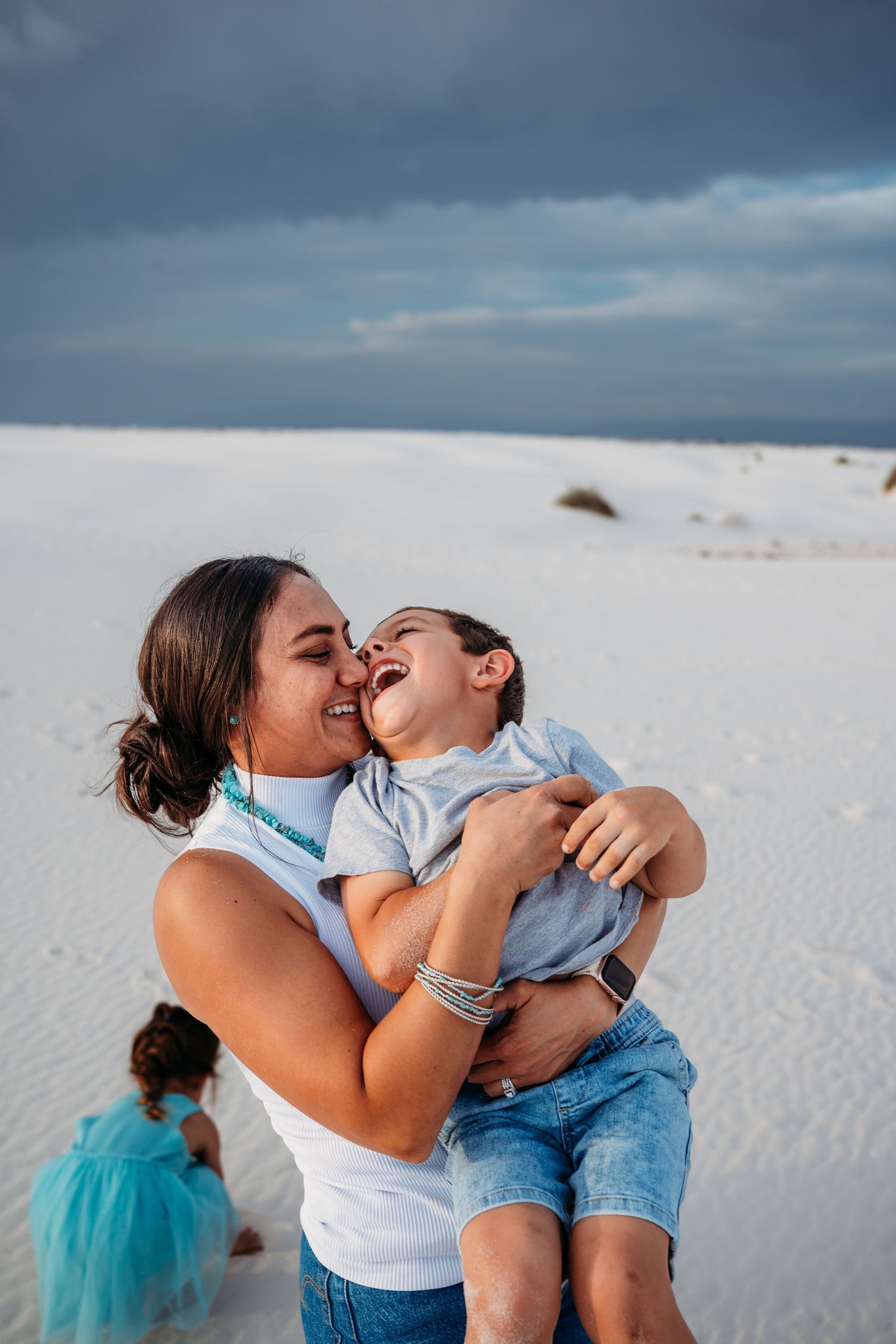A mother laughs joyfully while holding her young son on white sand dunes under a cloudy sky.