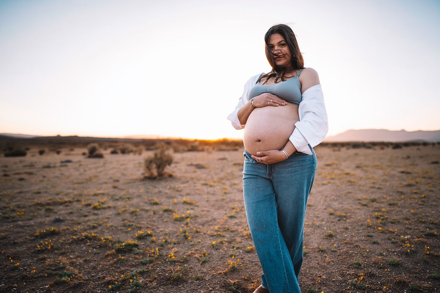 Pregnant woman with arms crossed over belly wearing jeans in desert at golden hour.