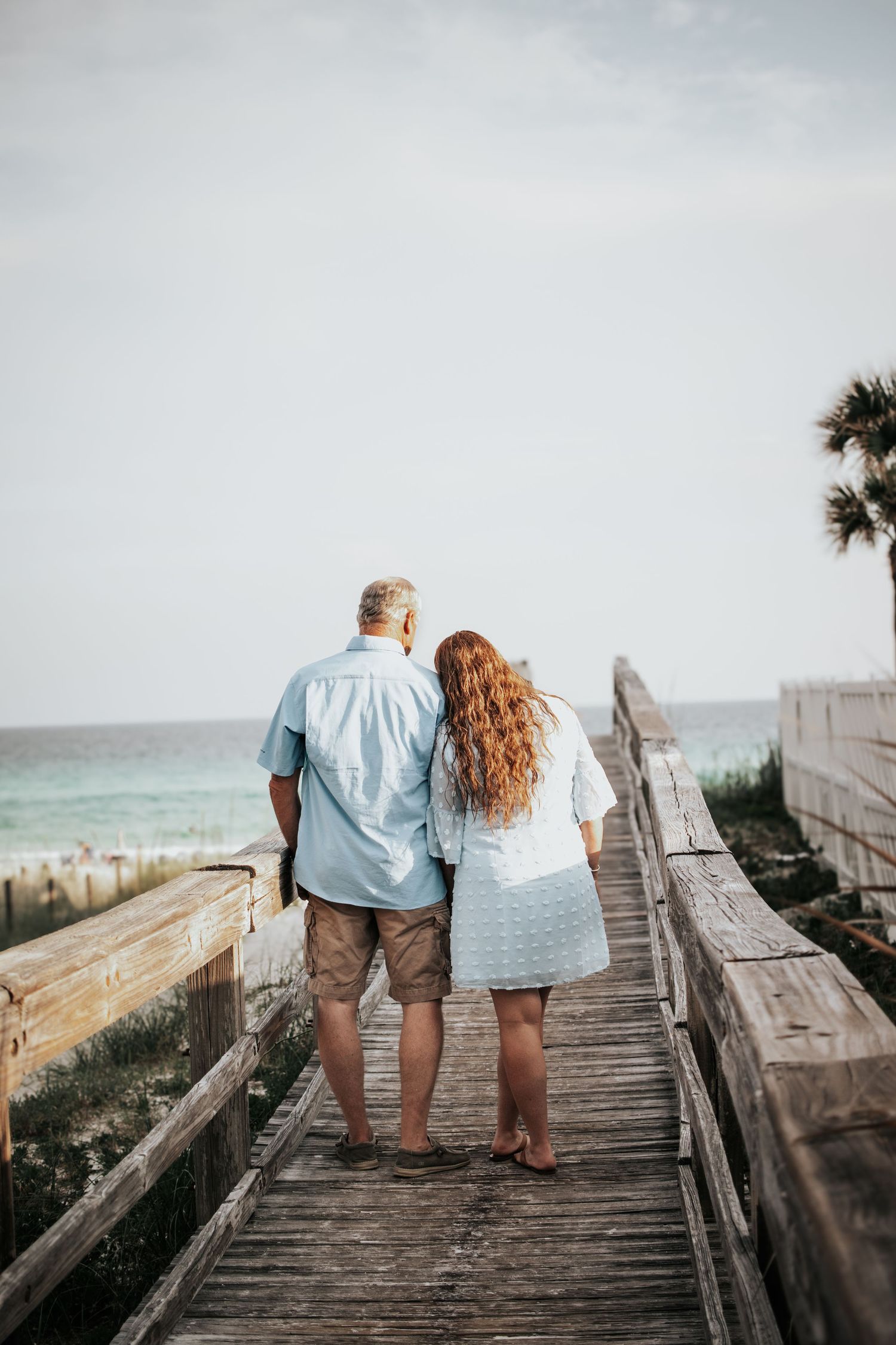 Mature couple walking hand-in-hand down a wooden boardwalk toward a beautiful turquoise beach.