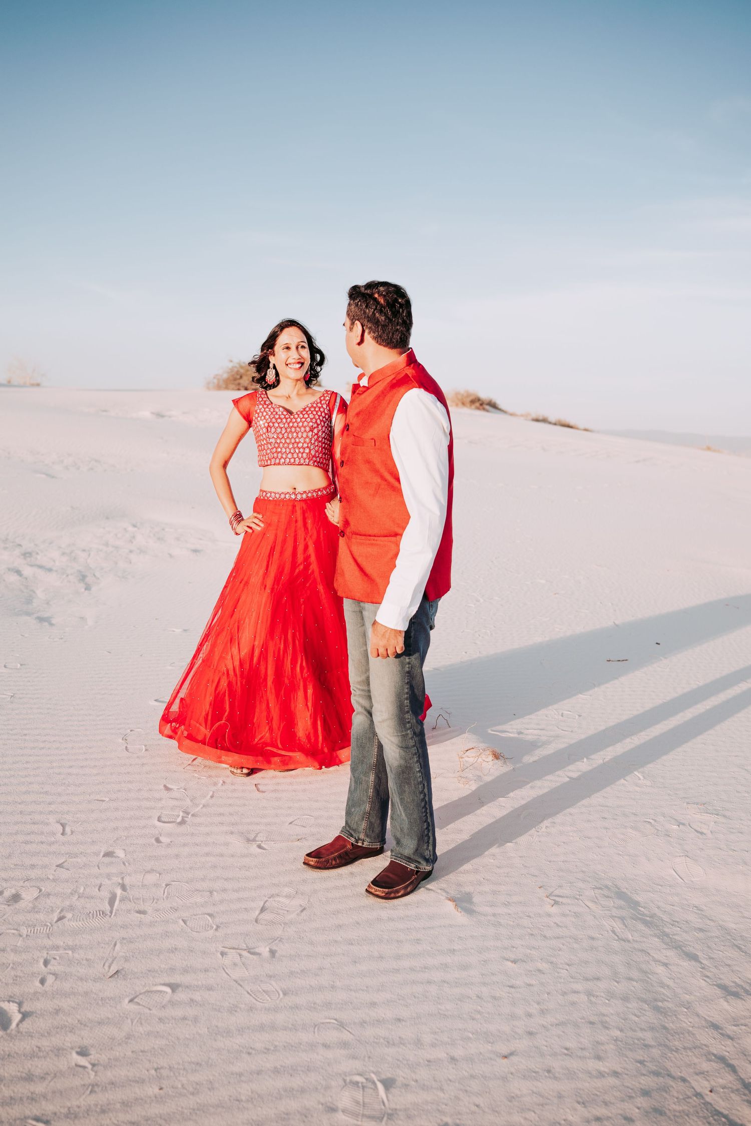 Indian couple in red outfits facing each other on white sand dunes under a clear blue sky.