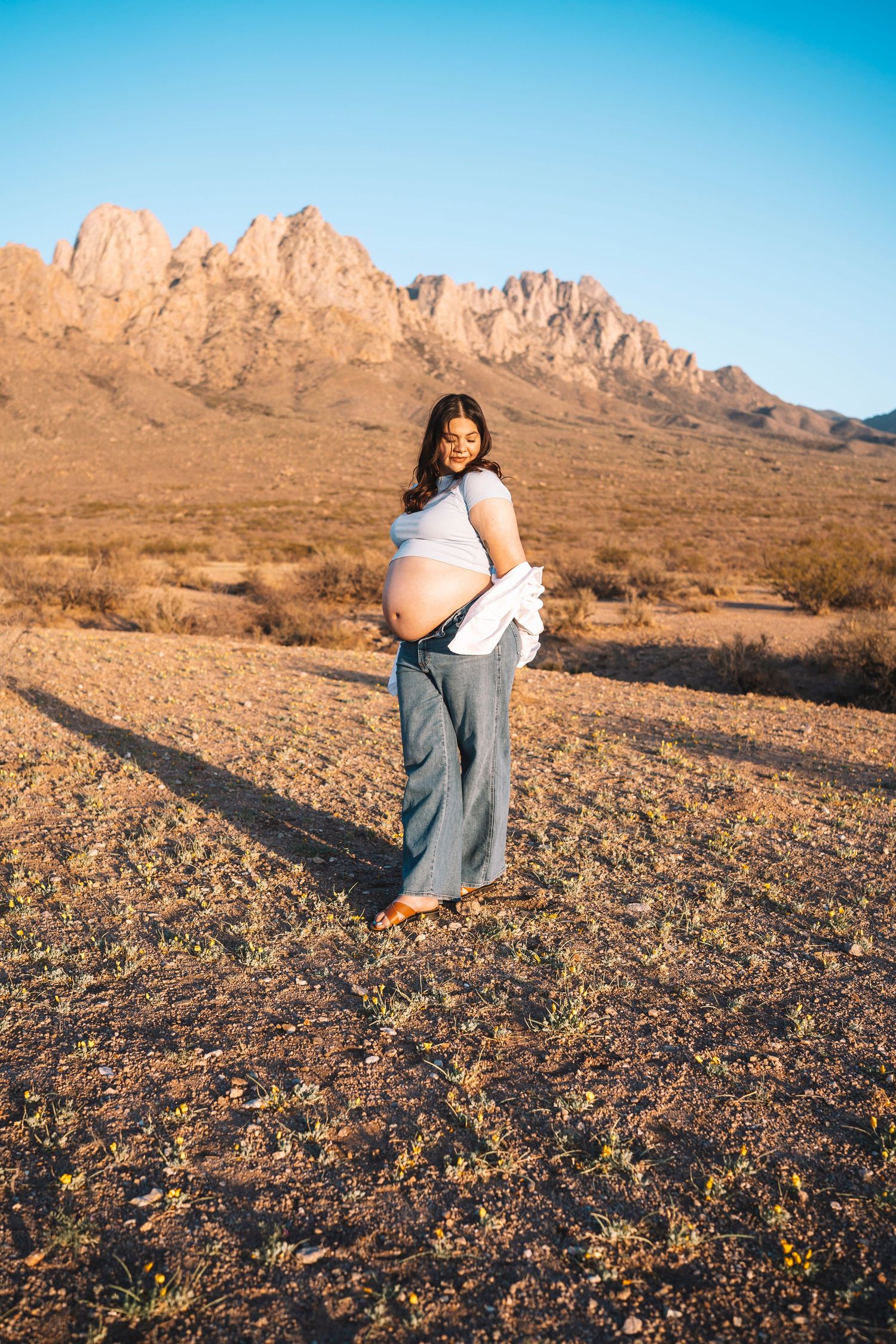 Pregnant woman in white top and jeans cradles baby bump in desert with mountain backdrop.