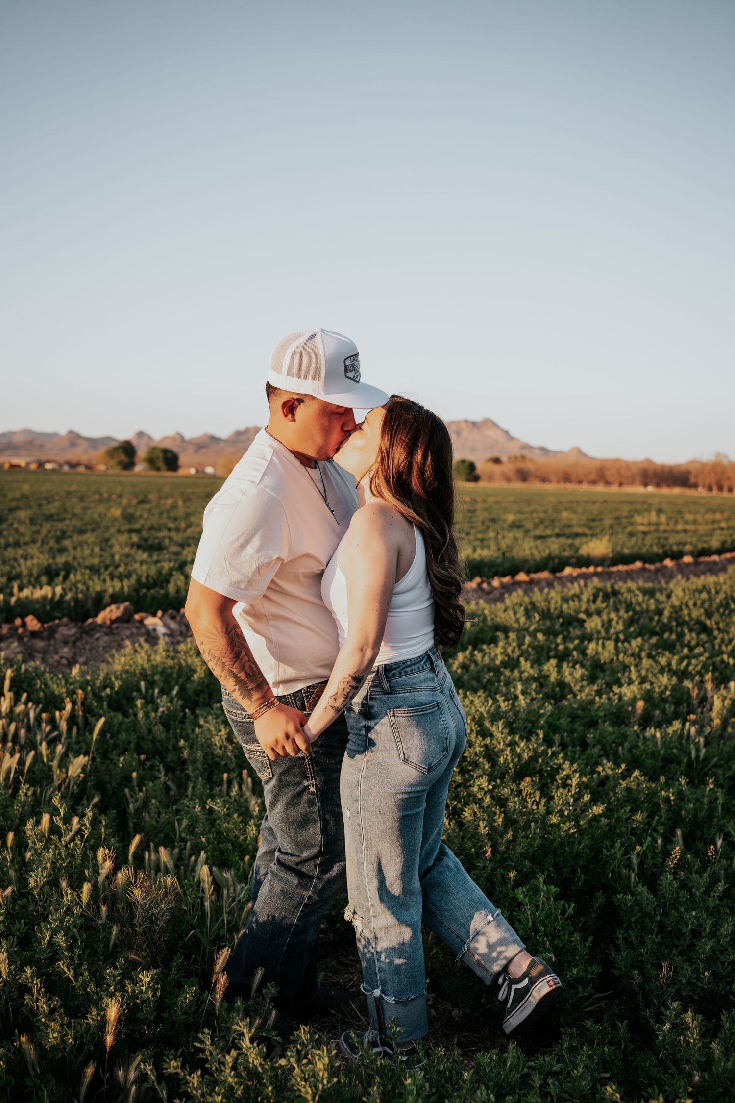 Couple sharing a kiss in a lush green field at sunset with mountains in the background.