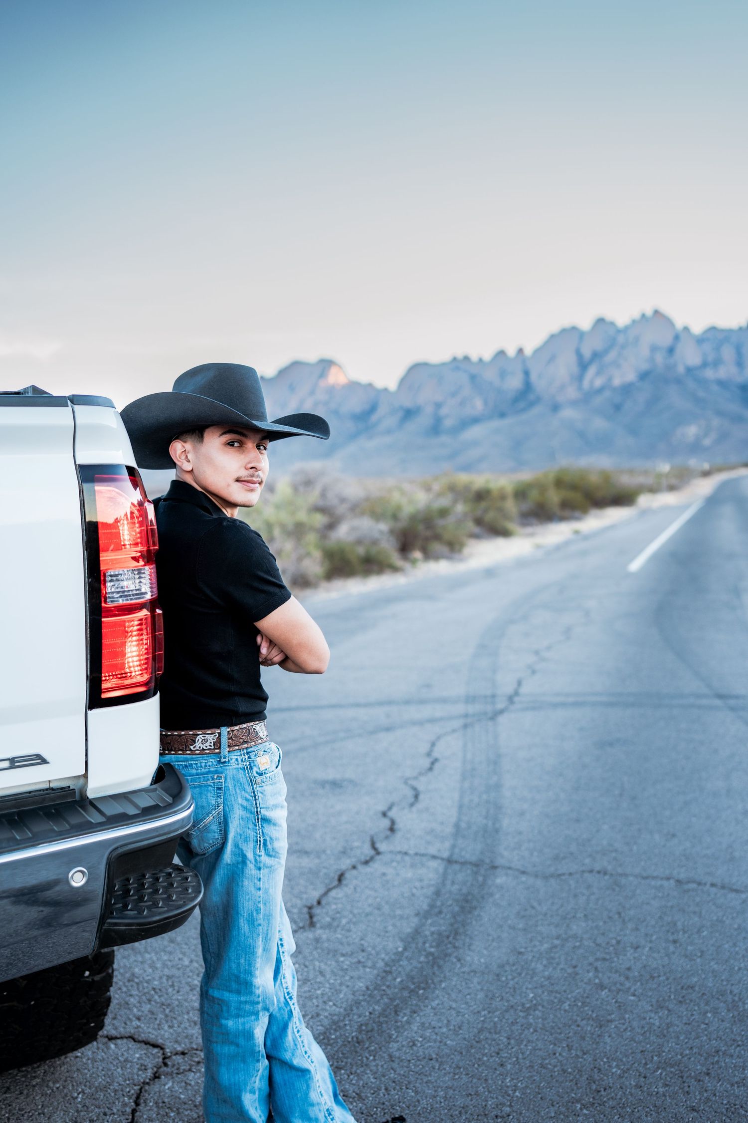 Young cowboy in black hat leans against white pickup truck on desert road with mountain scenery.