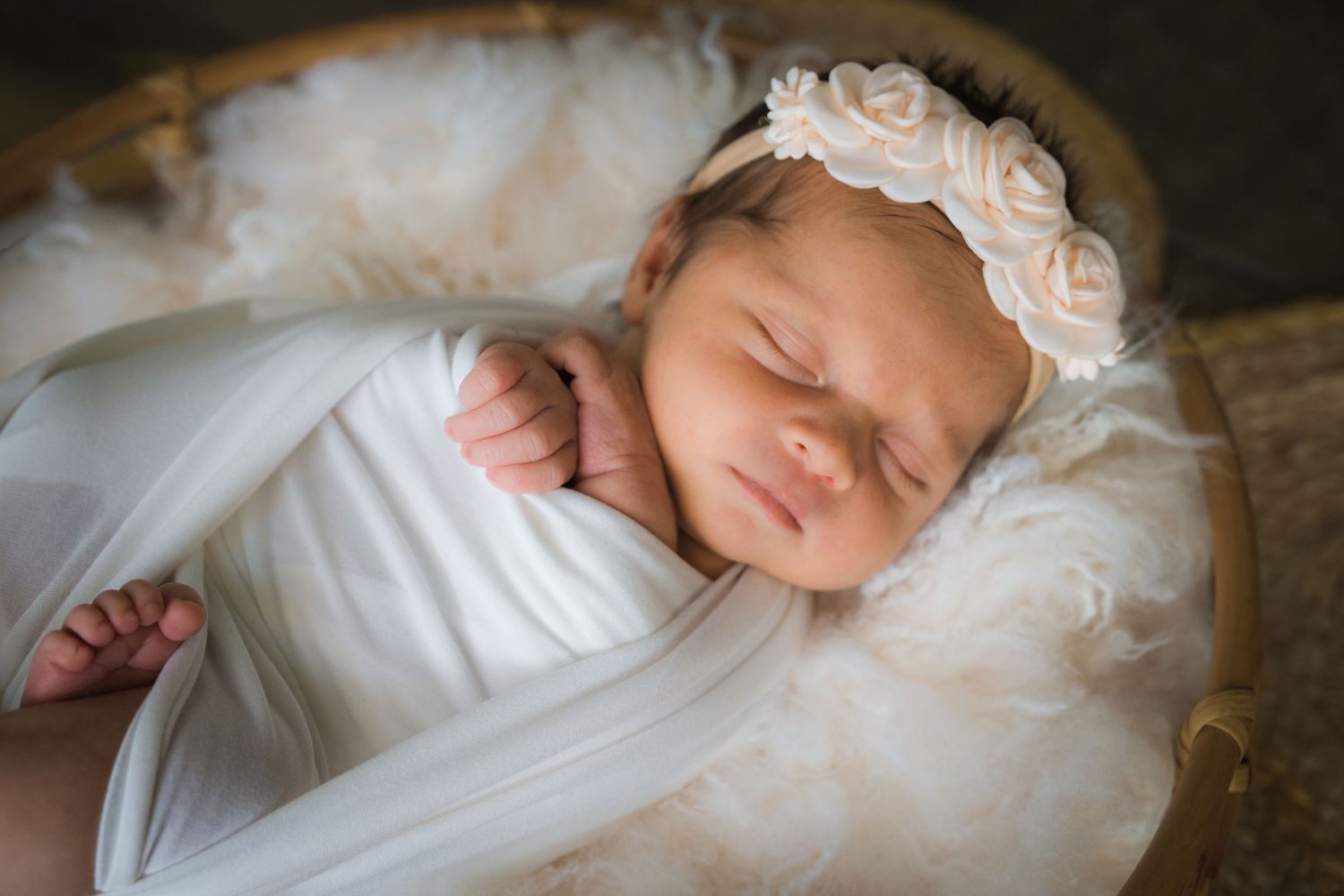 Peaceful newborn baby girl wrapped in white swaddle with rose headband sleeping in a wicker basket.