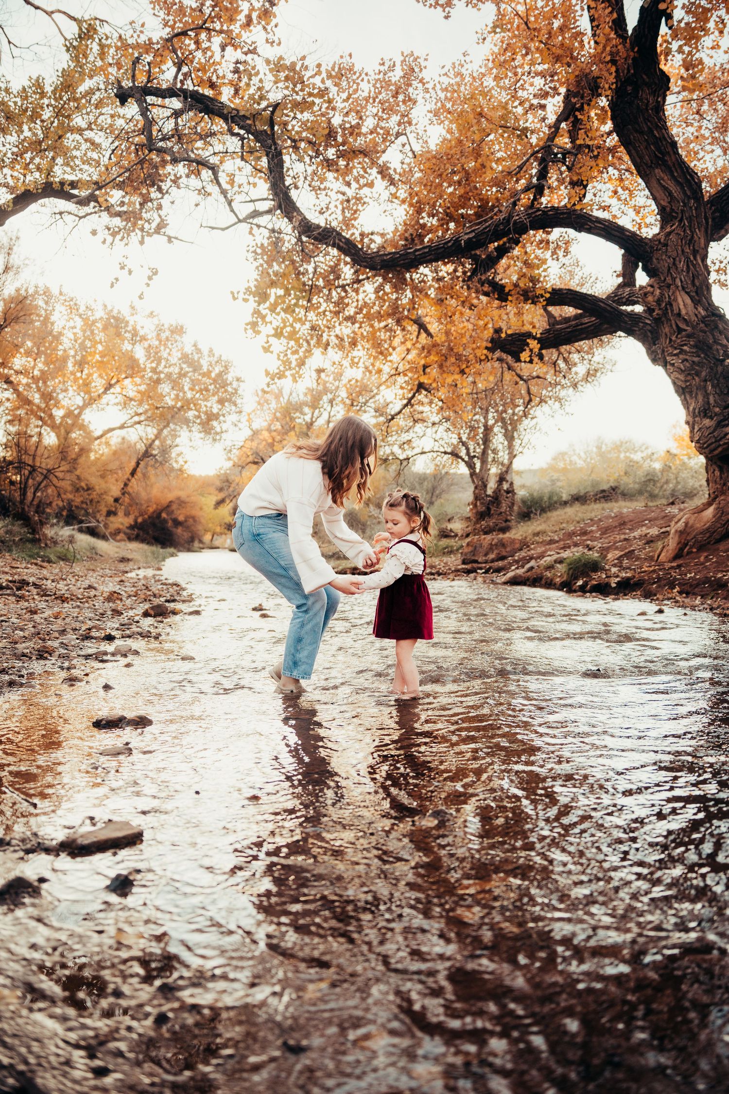 Mom bends down to interact with toddler daughter standing in autumn creek together.