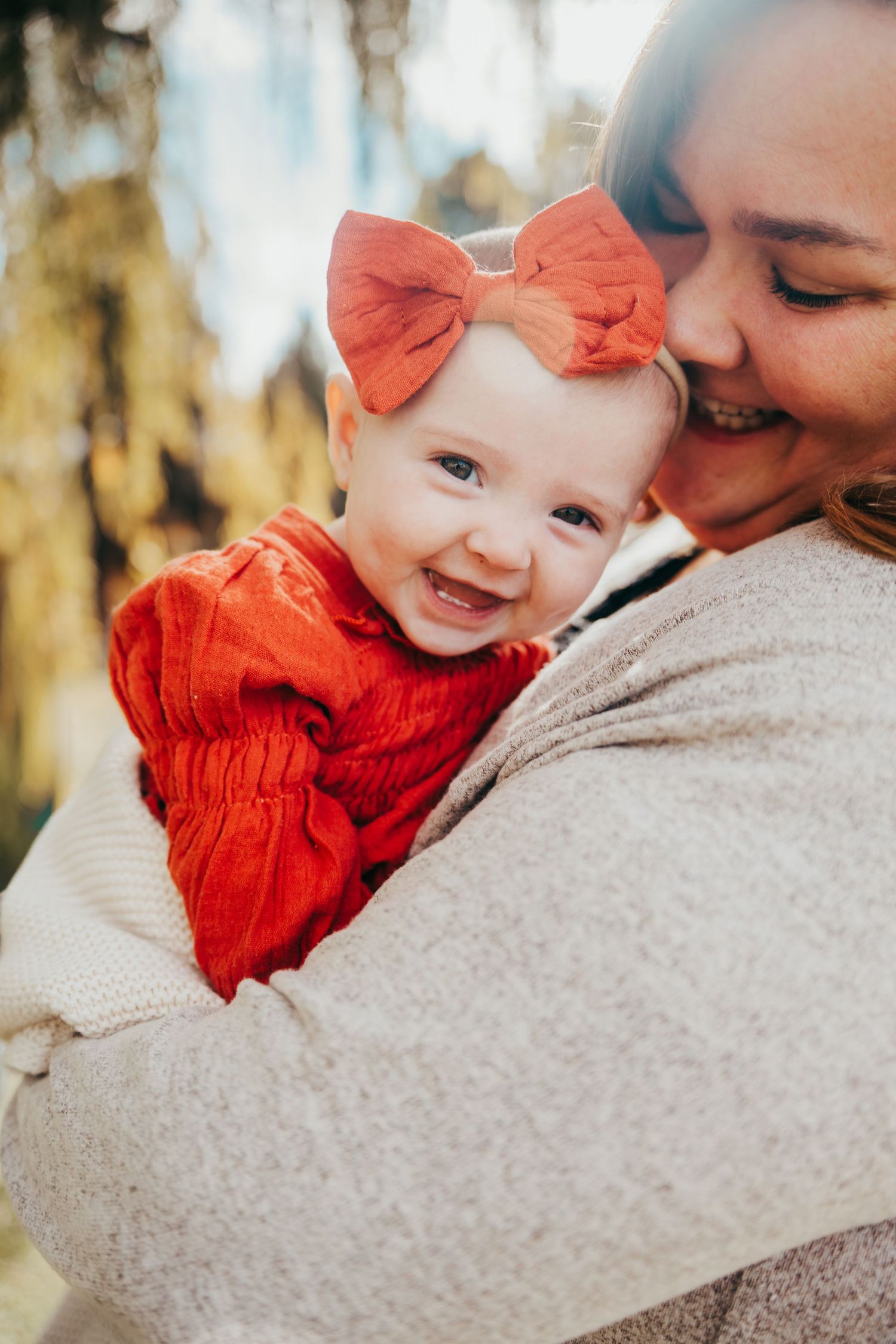 Mother kissing smiling baby girl in orange dress and red bow outdoors in autumn setting.